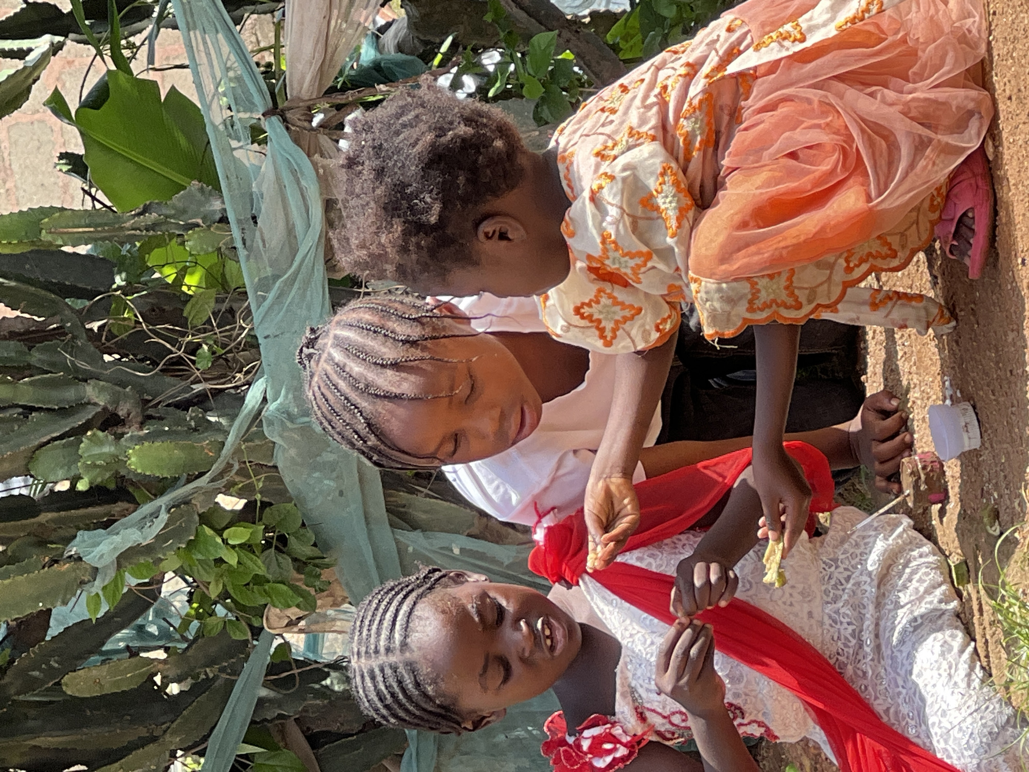 In Jos, Nigeria, three young girls are gathered outdoors, sitting on the ground near a large cactus plant. The girl on the left, wearing a white dress with red accents, holds a piece of food and appears to be eating. In the center, another girl with braided hair in a white shirt looks on, extending her hand with a piece of food, possibly offering it to the girl on the left. The youngest girl on the right, wearing a peach-colored dress with orange embroidery, is also present, looking towards the interaction. The ground is covered in dirt and small stones, with some scattered green leaves. A light green plastic sheet is draped over some of the vegetation in the background, and a brick wall is visible further back.