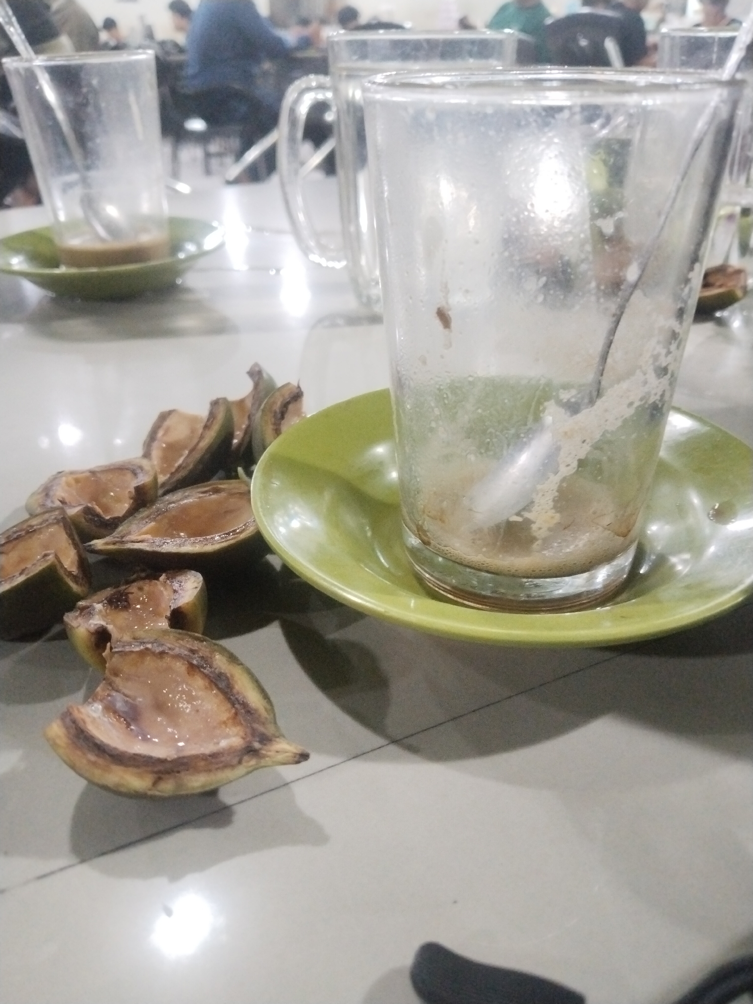 The image depicts a table setting at what appears to be a restaurant or coffee shop in Lhokseumawe, Indonesia. There are several empty glass cups on green saucers. One cup contains a spoon and residue of a beverage, likely coffee. Scattered around the table are several slices of fruit, potentially rambe. People are visible in the background, seated at other tables. The environment has a casual, public setting.