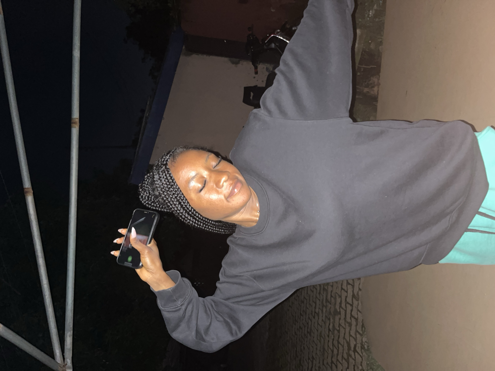 The image shows a Black woman standing outdoors at night, possibly in Jos, Nigeria, holding a smartphone in her right hand above her head. She is wearing a black crew neck sweatshirt and turquoise shorts. Her eyes are closed, and her head is tilted back, suggesting she might be enjoying music or posing for a photo. The background includes a brick-paved area, a building with a blue trim, and some vegetation. A motorcycle is partially visible in the background. The lighting suggests artificial illumination from an overhead source.