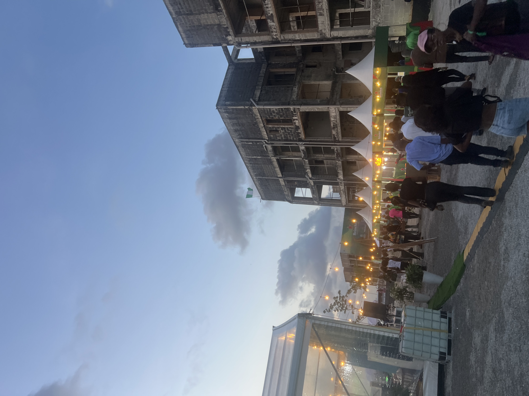 The image depicts an outdoor event held in Lagos, Nigeria, at dusk. An unfinished, multi-story concrete building serves as a backdrop to the event. A Nigerian flag is visible on the building. Several white tents line the event space, illuminated by string lights. People are seen standing and interacting, their faces largely obscured. A semi-transparent structure, possibly a tent or enclosure, is in the foreground. Heineken banners are visible. The sky is partly cloudy.