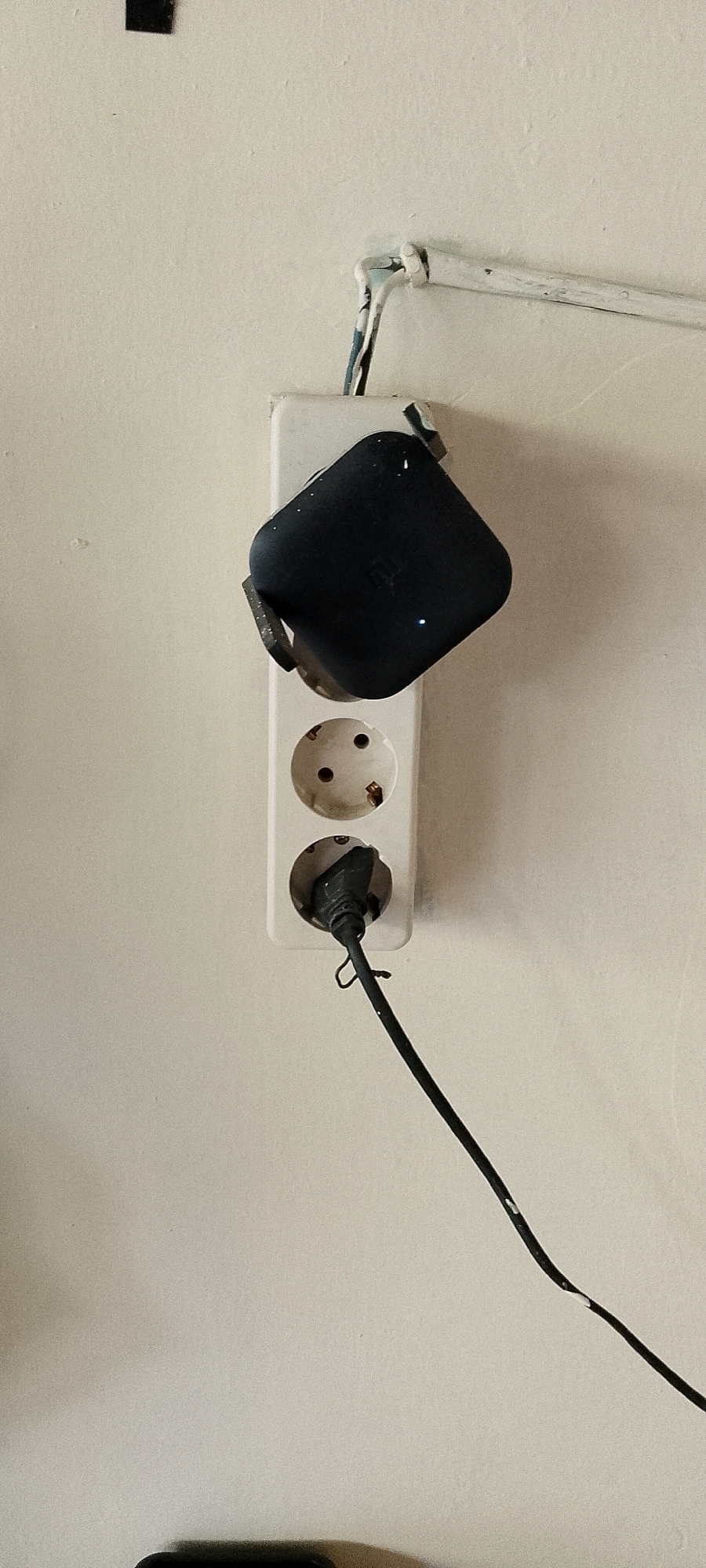 In Satriamekar, Indonesia, a close-up view captures an electrical outlet on a light-colored wall. A white, two-socket power strip is mounted vertically. A black charging brick, possibly for a mobile device, is plugged into the top socket. A blue indicator light is visible on the charging brick. A black power cord extends from the bottom socket and snakes downwards and to the right across the wall. The cord appears to have some minor damage or wear. Above the power strip, a white pipe runs horizontally, secured to the wall by a white fastener and a piece of tape. The overall scene is dimly lit, highlighting the texture of the wall.
