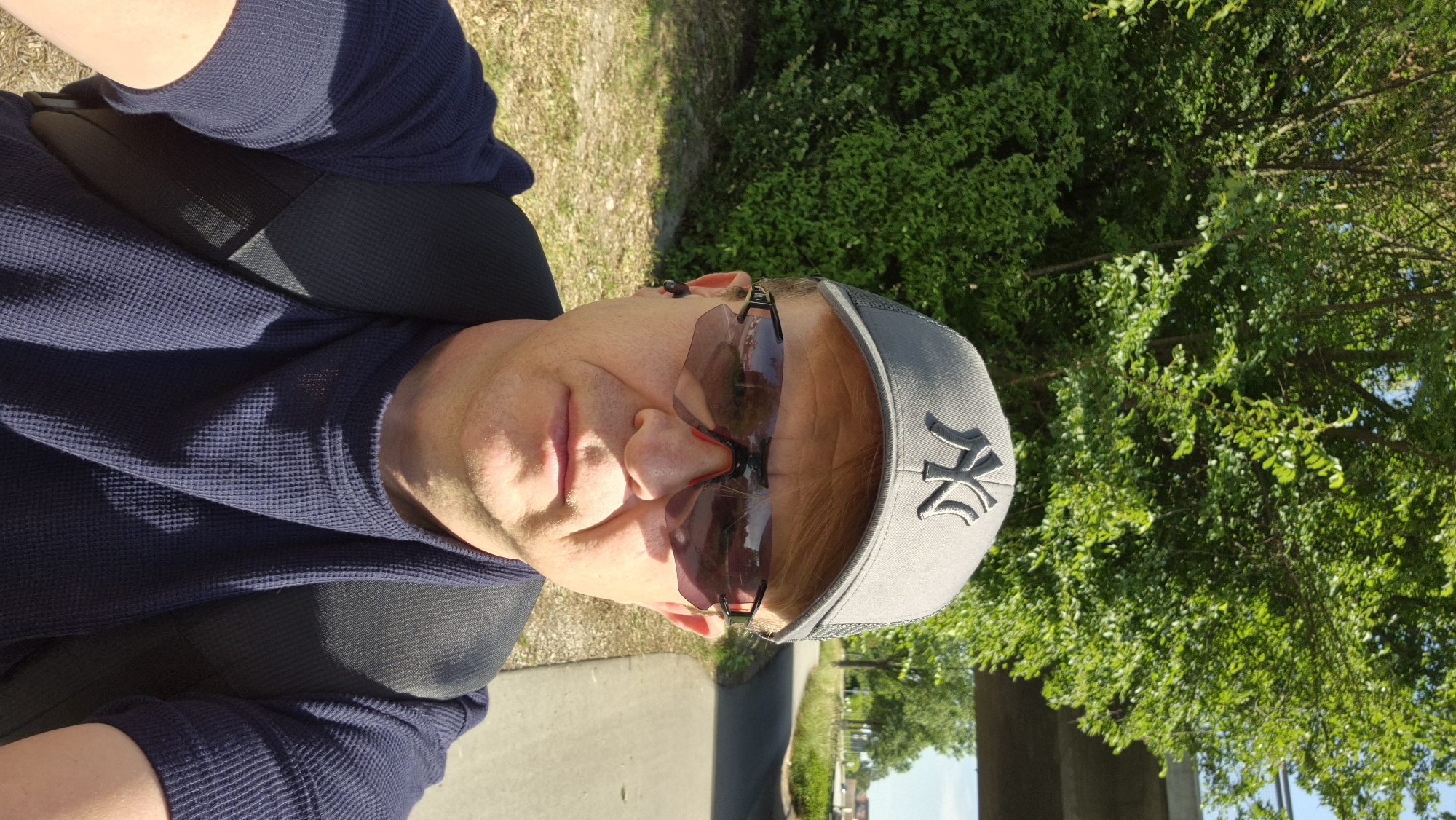 A male individual, appearing middle-aged, is captured in a sunlit selfie outdoors in the city of Unknown, Unknown. He wears a grey baseball cap with a dark "NY" logo, dark reflective sunglasses with red accents, and a dark blue short-sleeved waffle-knit shirt. A black earbud is visible in his left ear. He carries a black backpack, with the straps visible over his shoulders. The background features dense green foliage (trees and bushes) on the left, and a paved pathway extending into the distance on the right. A concrete overpass or bridge structure spans across the upper right portion of the image. The scene is brightly lit, indicative of daylight.