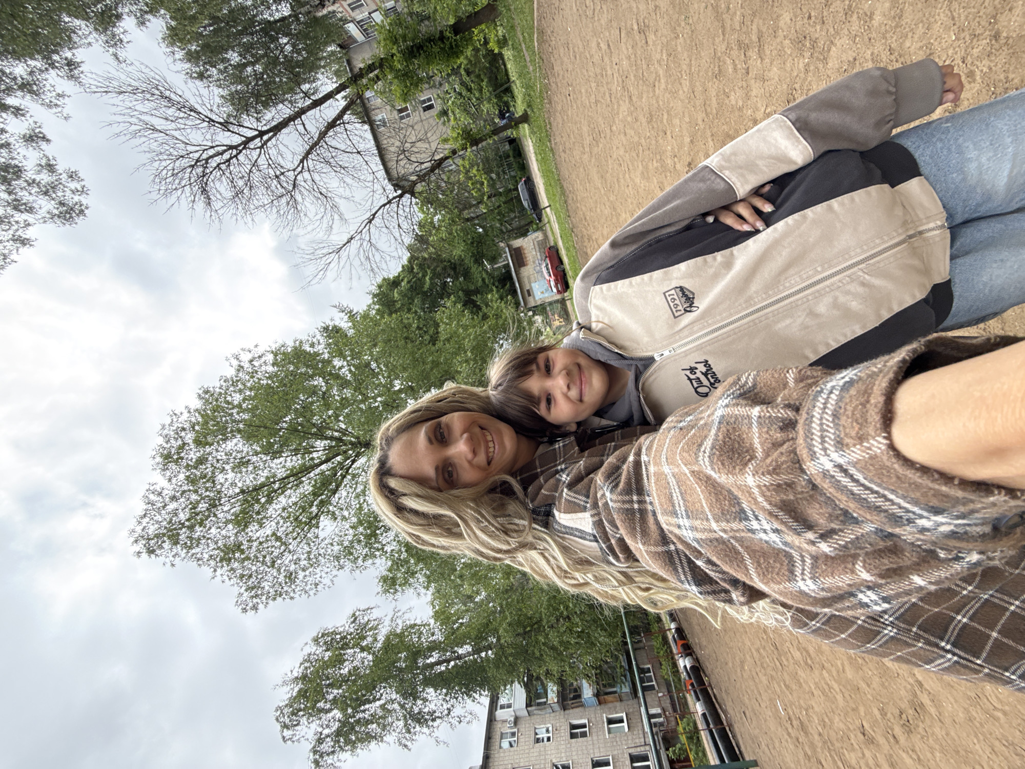 A photograph taken in Rusanovo, Russia, shows two people, one woman, and one child, posing for a selfie in an outdoor environment. The woman is in the foreground wearing a brown plaid shirt with long blonde curly hair. The child, assumed to be her child, is leaning against the woman. The child is wearing a grey jacket with black and beige accents. They are positioned on a dirt-covered ground. In the background, there are trees, buildings, and a red car. The sky is overcast.
