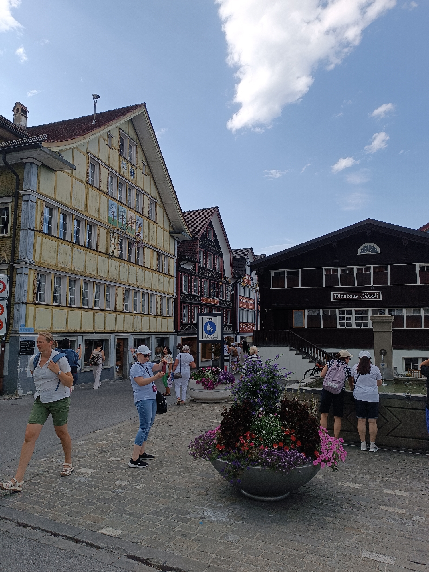 The image depicts a street scene in the city of Appenzell, Switzerland, likely on a sunny day given the bright lighting and visible clouds. In the foreground, a woman in a white shirt and green shorts walks from left to right, carrying a blue backpack. To her right, another woman in a blue shirt and jeans stands and appears to be taking a photo with her phone. A large, decorative planter filled with colorful flowers is situated between them and a fountain. Several other people are visible in the background, walking or standing near buildings and the fountain. The buildings themselves are characteristic of Swiss architecture, with traditional timber-framed facades and steep roofs. One building on the right has a sign that reads "Wirtshaus Rössl". The street is paved with cobblestones.