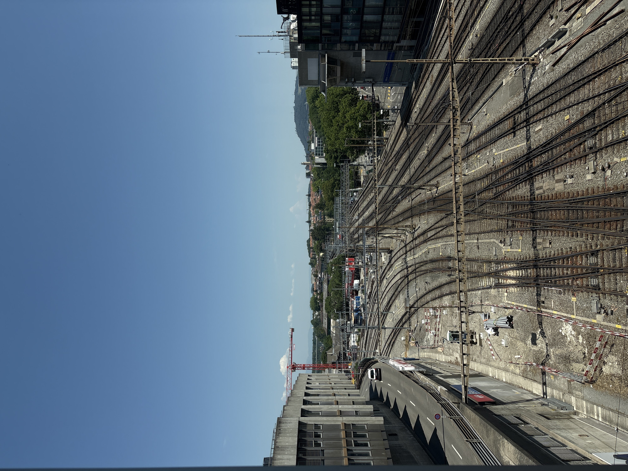 The image depicts a wide shot of a train yard in Bern, Switzerland, on a clear, sunny day. In the foreground, a concrete building with rows of windows lines the left side of the frame, with a curved road running alongside it. A white van is visible on the road.
Dominating the middle ground is an extensive network of train tracks, curving and branching in various directions. A red construction crane stands prominently in the distance, to the left of the tracks. Beyond the train yard, a dense canopy of green trees forms a backdrop, with buildings and structures interspersed amongst them. The distant skyline shows more buildings and a hazy mountain range under a bright blue sky with a few scattered clouds.
The scene suggests an active railway hub, possibly undergoing some construction or maintenance, given the presence of the crane and visible construction materials near the tracks in the lower center. The overall environment is urban, with a clear emphasis on the infrastructure of a railway system.