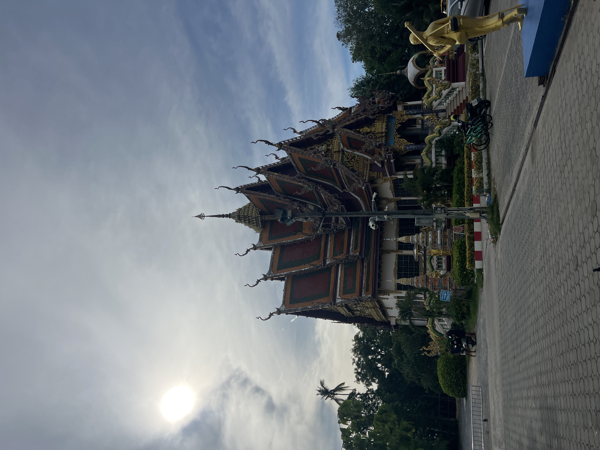 In Baan Plai Laem, Thailand, a golden statue of a man carrying a staff stands to the right of the frame, facing left. Behind the statue, a temple with ornate, multi-tiered roofs and intricate carvings is visible. The temple has a white facade with colorful accents. To the left of the temple, lush green trees and a palm tree create a natural backdrop. The ground in the foreground is paved with gray interlocking bricks. A few motorcycles and bicycles are parked in front of the temple. Two individuals are standing near a motorcycle to the left of the temple entrance. The sky above is partly cloudy with a bright sun visible on the left side of the frame.