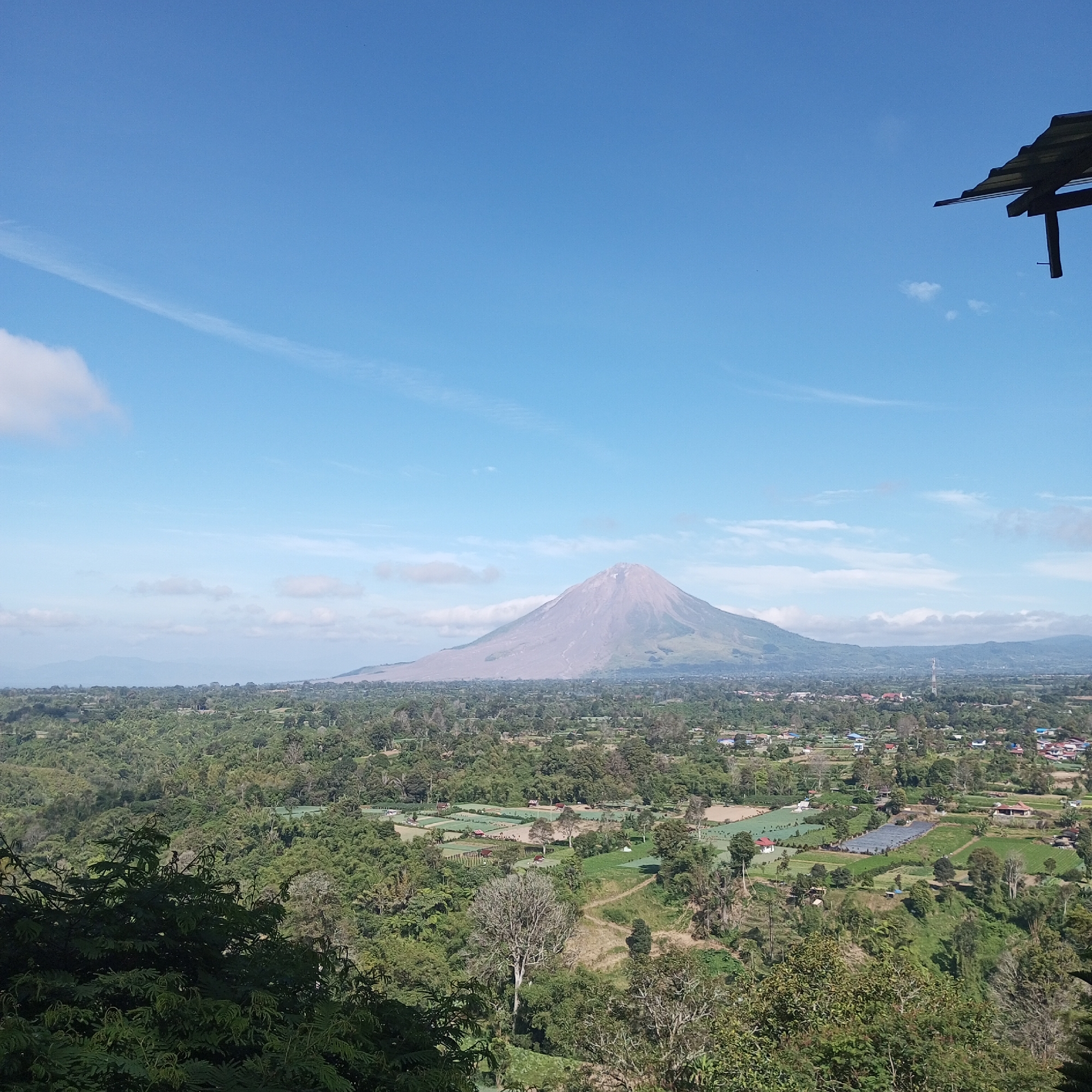 The image depicts a wide, scenic landscape viewed from an elevated vantage point. The sky is a clear, vibrant blue with sparse white clouds. Dominating the background is a large, conical mountain with a light-colored, possibly ash-covered peak, suggesting volcanic activity. Below the mountain, a sprawling green landscape unfolds, featuring lush vegetation, trees, and hints of agricultural fields or terraced land. Scattered throughout the middle ground are buildings, some appearing to be homes or small structures, and a visible cell tower. In the foreground, dense foliage and trees create a natural frame for the view. The overall scene suggests a rural or semi-rural environment with a prominent natural landmark. The location is identified as Gundaling 2, Indonesia.