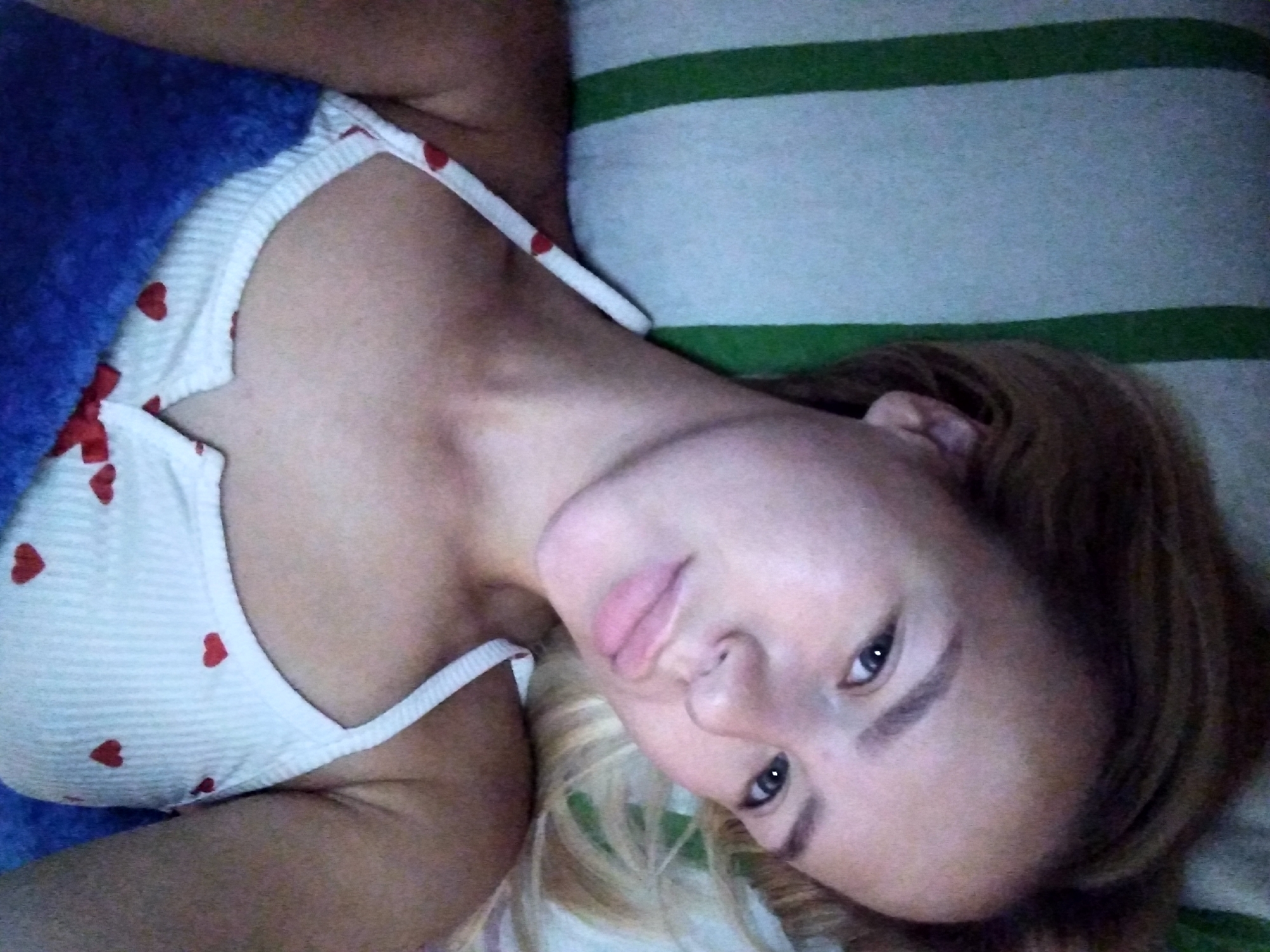 A young woman with blonde hair and fair skin is lying on a bed, looking at the camera. She is wearing a white tank top with red hearts on it, and a blue towel is draped over her. The background is a striped green and white pillow. The scene appears to be in a bedroom in the city of Radisne, Ukraine.