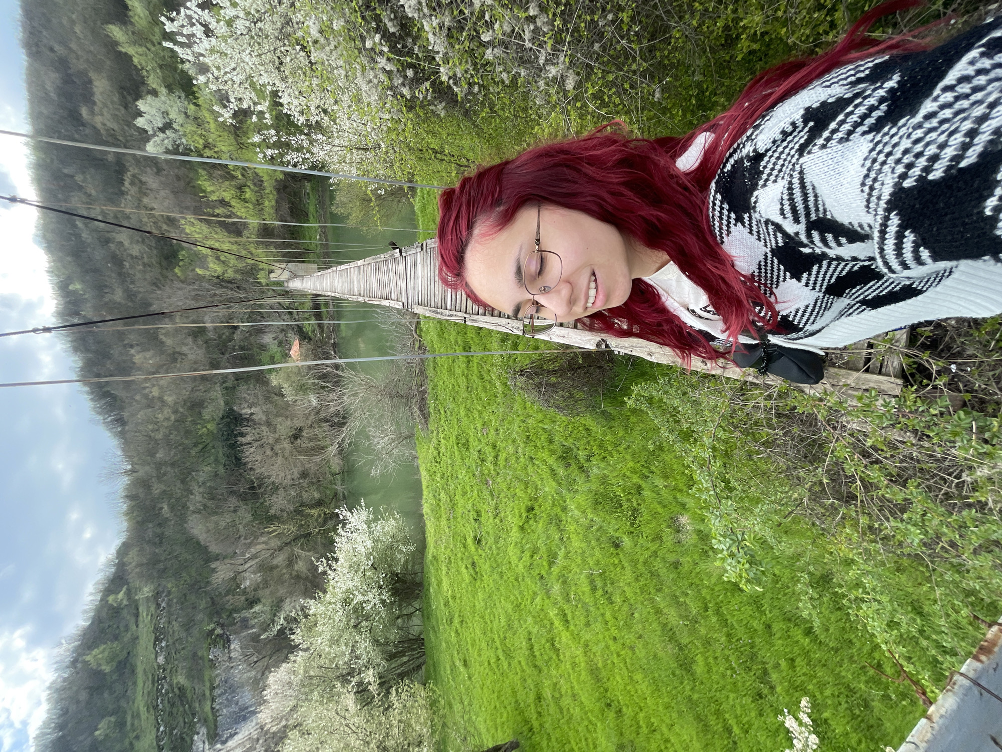 A young woman with red hair and glasses is standing on the edge of a suspension bridge in Sadovets, Bulgaria. She is wearing a black and white patterned sweater. Behind her, the bridge stretches over a river, and the surrounding landscape is lush with greenery and trees, some with white blossoms. The sky is overcast.