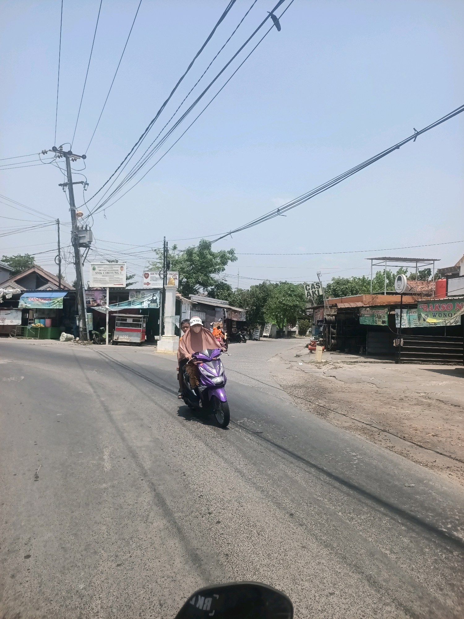 In Wanajaya, Indonesia, on a clear day, two individuals ride a purple scooter down a paved road. The scooter is in motion, heading away from the viewer. The person in the front, wearing a light brown hijab and a white cap, is driving. The person in the back, a child, is also wearing a light brown hijab. They are riding on the right side of the road.
The road is flanked by a sidewalk and a line of buildings on the left, and a dirt shoulder and more buildings on the right. Several shops are visible, including one with a sign that reads "BAKSO N WONOGIRI" and another with "CAS AKI". Overhead, numerous power lines crisscross the sky, supported by utility poles. Trees with green foliage are scattered throughout the scene, suggesting a suburban or semi-urban environment. The sky is bright blue with a few faint clouds. The overall impression is a snapshot of daily life on a road in Indonesia.