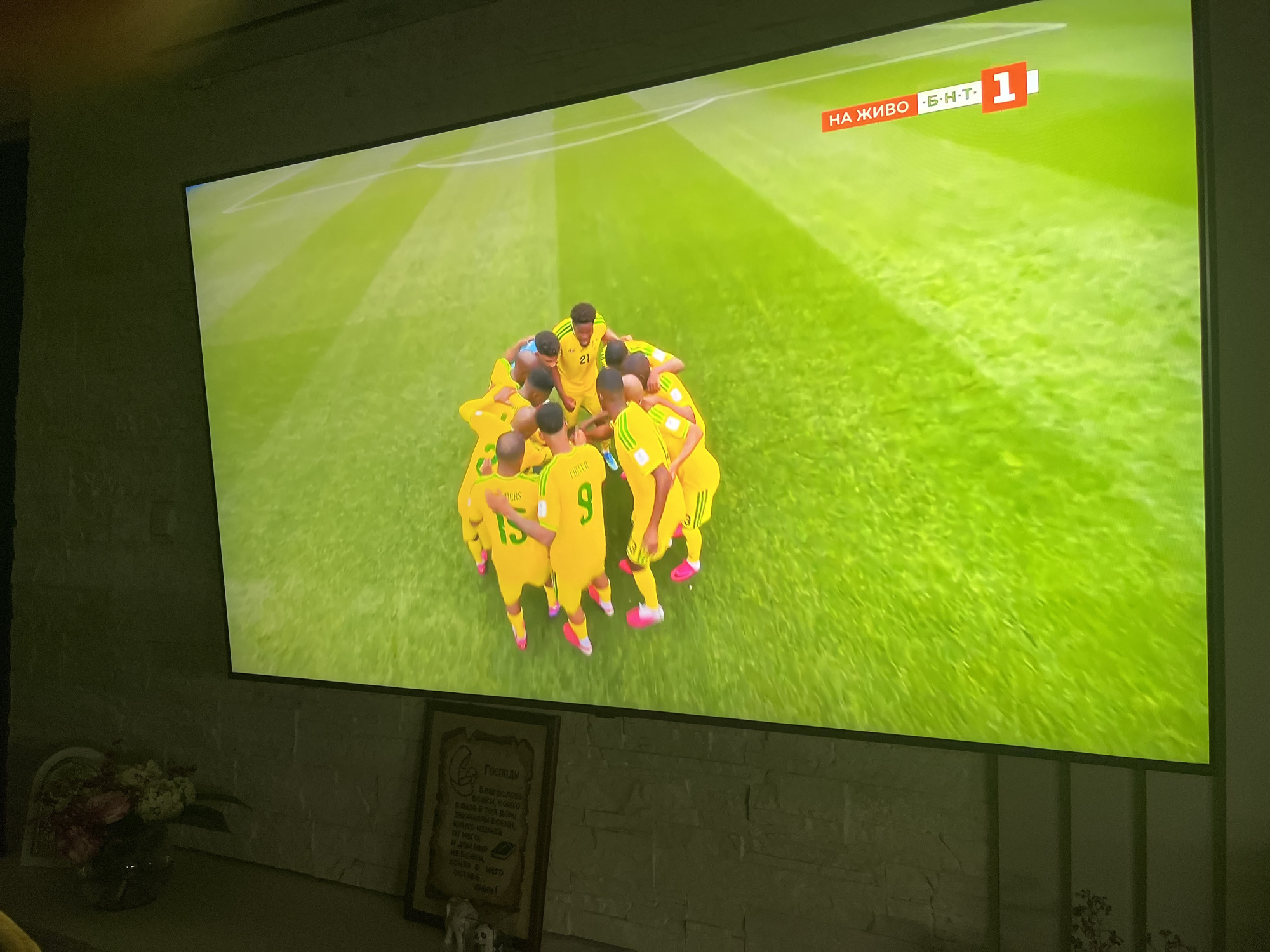 The image captures a television screen displaying a live broadcast of a football match, likely from Varna, Bulgaria, given the context. The screen shows a huddle of players in yellow jerseys with green accents and white numbers. Several players have numbers visible on their backs, including "15" and "9." The players are positioned on a vibrant green football field, possibly with white lines marking the boundaries. In the upper right corner of the screen, there is a broadcast graphic that reads "НА ЖИВО" (which means "live" in Bulgarian) and "B.H.T. 1", suggesting the channel is BNT 1.
Below the television, a brick wall serves as a backdrop. On a surface in front of the wall, a vase with flowers is visible, along with a framed picture or document. The lighting in the room is dim, with the primary illumination coming from the television screen, casting a green hue onto the surrounding environment. The overall scene appears to be a living room or entertainment area where the football match is being watched.