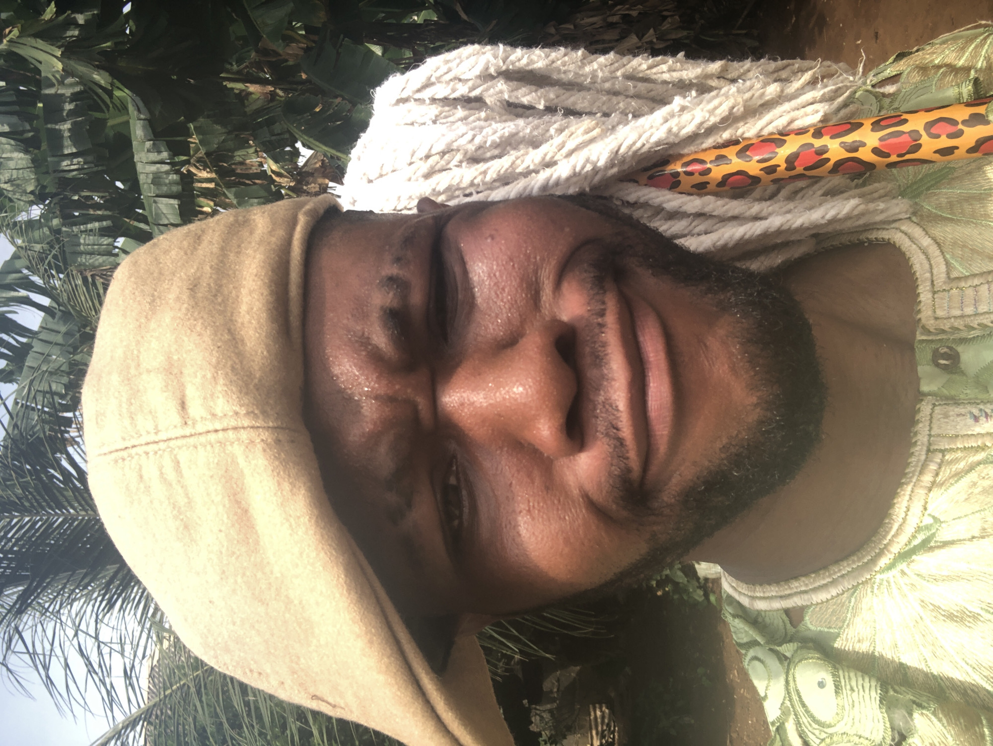The image, oriented correctly, features a close-up portrait of a man against a backdrop of lush green foliage. The man has dark skin, a short beard, and a mustache, and is looking directly at the viewer with a relaxed expression and a slight smirk or smile. His face appears shiny, possibly from sweat or oil.
He wears a light beige or tan hood or head covering that frames his face. Over his shoulders and chest, he wears a light green garment, possibly a traditional robe or kaftan, adorned with intricate, shiny embroidery or patterned fabric featuring circular motifs.
Behind and slightly above his head is a white, rope-like mop head. A distinctive mop handle, featuring a bright orange base with a leopard print pattern (black and red spots), extends diagonally from the mop head across the upper right side of the frame.
The background consists of dense, vibrant green leaves, suggesting tropical or subtropical plants like banana leaves and palm fronds. The lighting is bright and natural, indicative of daylight. The overall scene conveys a casual, perhaps playful, moment outdoors, blending traditional attire with an everyday object. No text is visible in the image.