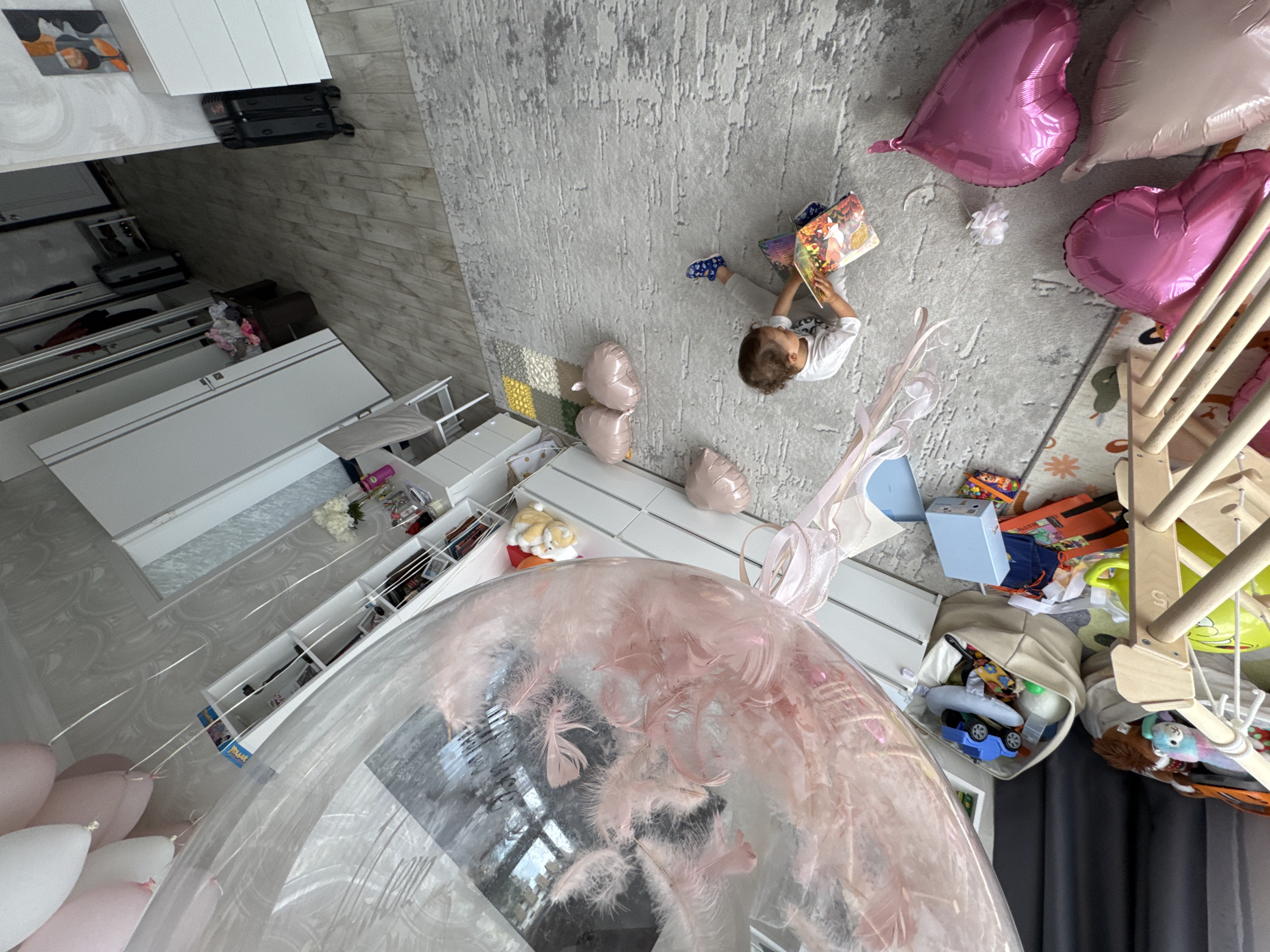 A high-angle shot captures an interior scene, likely in Sochi, Russia, considering the possible origin of the image. A young child is seated on a light gray patterned rug, absorbed in reading a book. Near the child are several heart-shaped pink balloons. The room includes a wooden climbing frame, toys, and assorted objects. The room also contains white furniture and what appears to be a large decorative balloon filled with pink feathers.