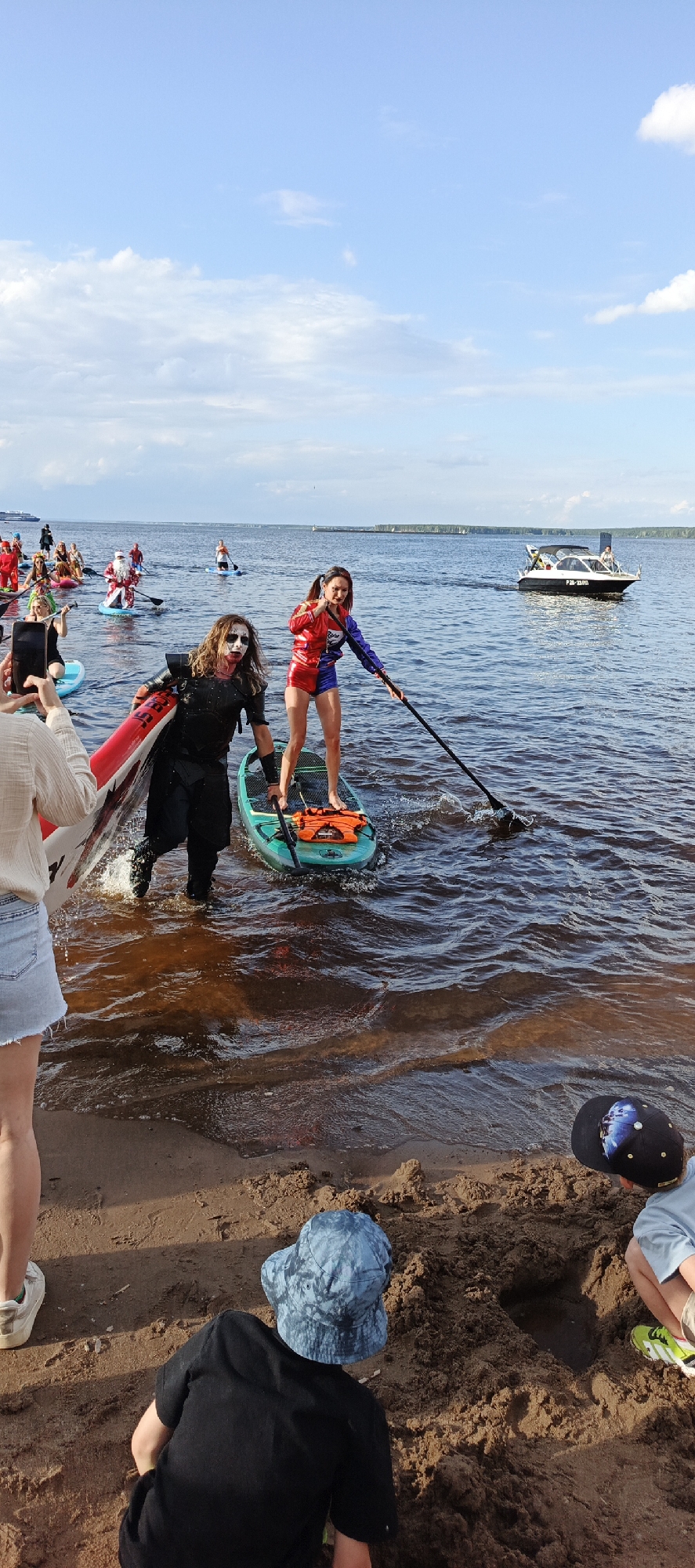 In the city of Chaikovsky, Russia, on the Kama River, a group of people are paddleboarding. The sky is partly cloudy, with scattered white clouds against a blue background. In the foreground, on the sandy bank of the river, two children are partially visible. One child, wearing a blue patterned bucket hat and a black t-shirt, is facing away from the camera, appearing to dig in the sand. Another child, wearing a light blue t-shirt and a black baseball cap, is also partially visible, with their back to the camera.
On the water, two individuals are prominently featured on paddleboards. One person, dressed in a black outfit with white face paint, is holding a red and white paddleboard. They appear to be stepping out of the water onto the shore or a shallow area, with their legs submerged. Behind them, on another paddleboard, a person dressed in a red, blue, and yellow superhero-like costume stands and holds a paddle, actively engaged in paddling. This individual has dark hair tied in pigtails.
Further in the background, a flotilla of paddleboarders can be seen, many in costumes. Some are dressed as Santa Claus, others appear to be in various outfits, suggesting a themed event. A white motorboat with several people on board is moving away from the shore in the middle ground. The water is calm, with small ripples, and the Kama River stretches towards a distant treeline. The overall scene depicts an outdoor event or gathering on the water.
