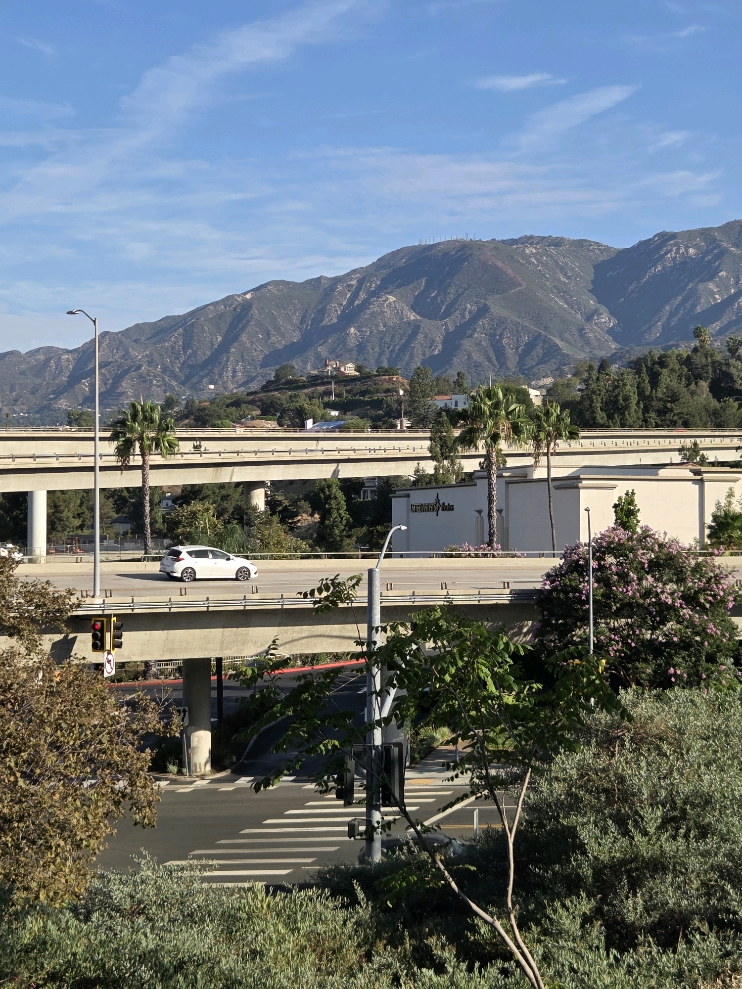 The image captures a view of the Glendale 210 Freeway in Glendale, California, with a backdrop of the San Gabriel Mountains. In the foreground, a white car drives on an elevated highway overpass. Below, another highway overpass is visible, with a traffic light showing red and a pedestrian crossing. Lush green trees and bushes surround the roadways. In the distance, the majestic San Gabriel Mountains rise, their slopes covered in vegetation and dotted with houses. The sky is a clear blue with a few wispy clouds. The scene conveys a sense of urban infrastructure harmoniously coexisting with natural beauty.