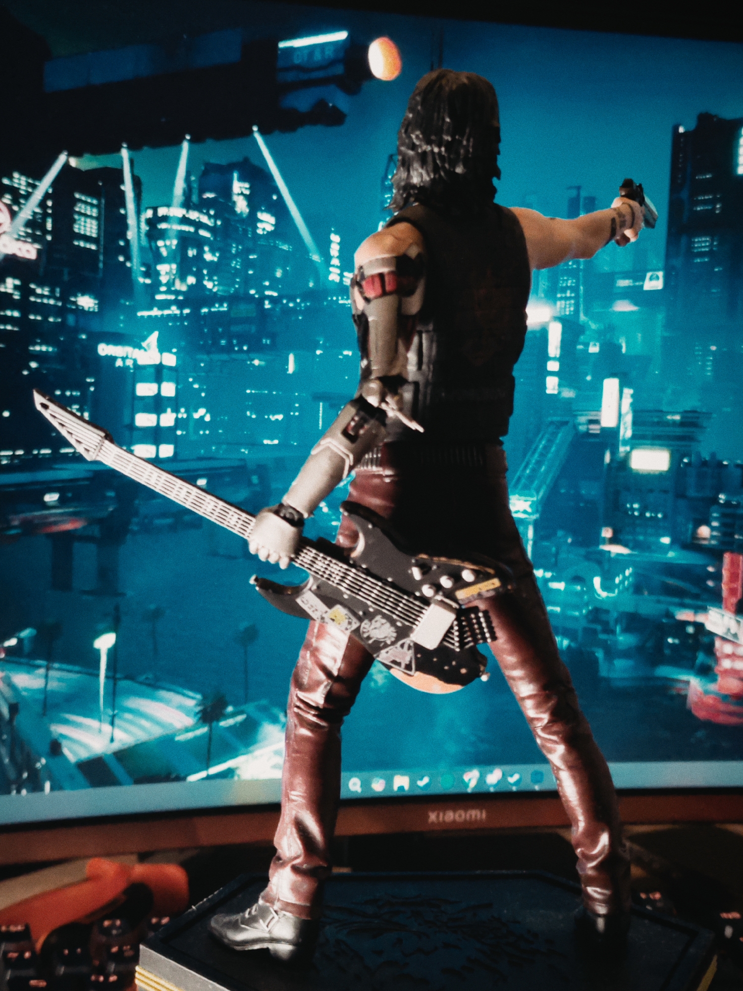 A figure of Johnny Silverhand from Cyberpunk 2077 stands with his back to the viewer. He is holding a guitar in his left hand and a gun in his right. The backdrop behind the figure is a brightly lit cityscape that appears to be from the video game Cyberpunk 2077. The figure is standing on a black platform with a gold trim. The scene is illuminated. A Xiaomi logo is visible. The environment does not show any element that would link this to the city of Budva, Montenegro.