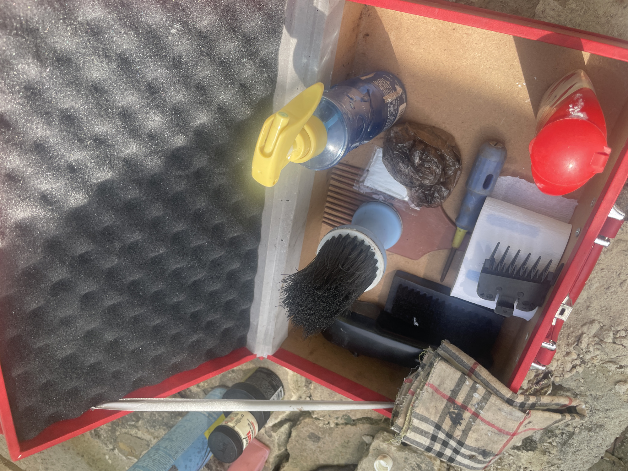 The image shows a red toolbox filled with various items resting on a rough outdoor surface. Inside the toolbox is a black foam pad, a clear plastic bottle with a yellow spray nozzle, a small wrapped dark object, a wooden comb, a light blue brush, a blue-handled tool, a roll of paper towels, a black comb attachment, a small black rectangular object, and a plaid cloth. On the surface beside the toolbox is a metal rod and other bottles. The location appears to be outdoors, judging by the rough surface and natural light. No specific city can be determined from this image.