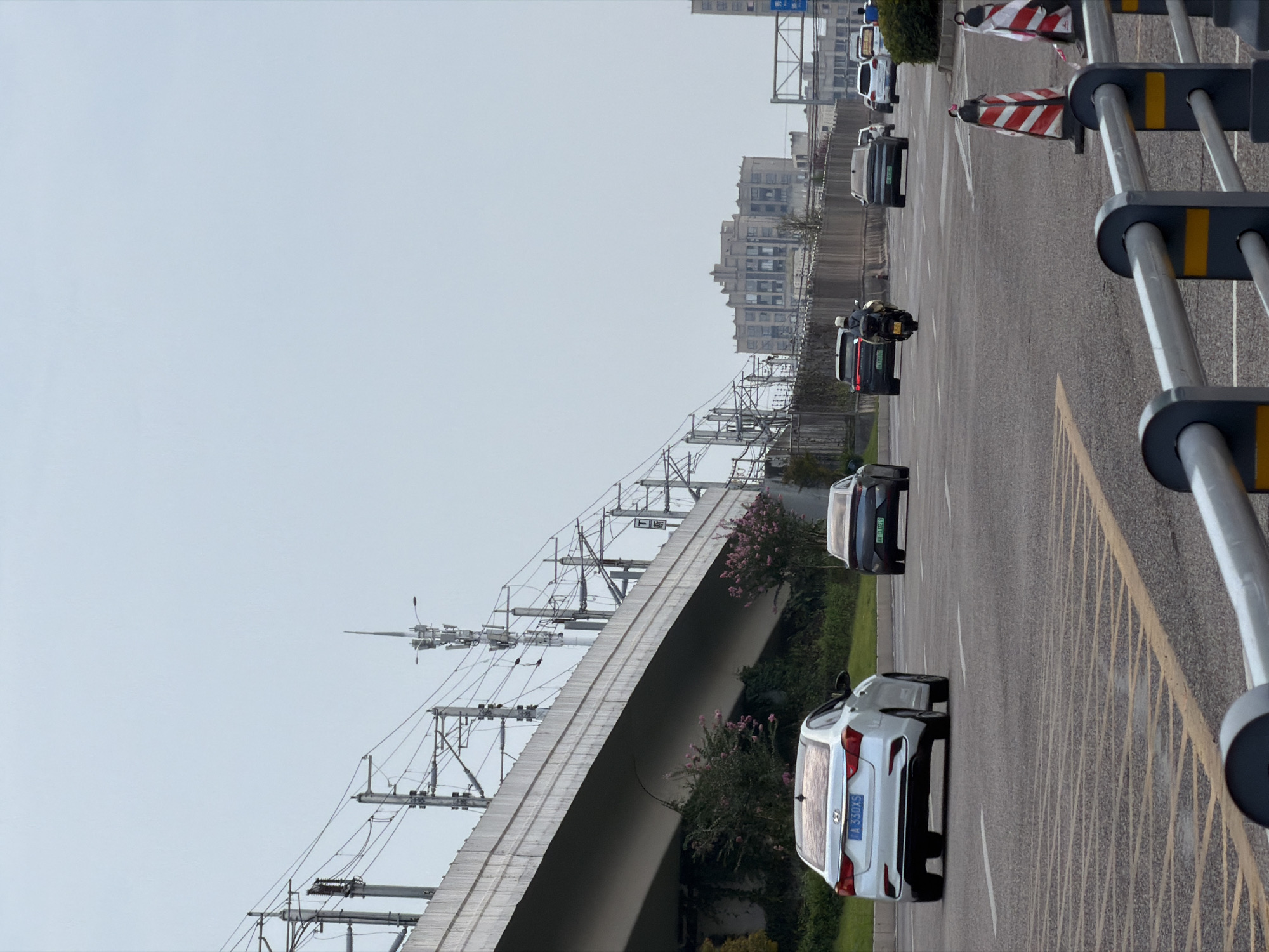 This is an eye-level, medium shot taken from a road in Jiangjin, China, depicting urban traffic and infrastructure. In the foreground, a gray metal guardrail with yellow markings separates the viewpoint from the road. The road itself is paved with asphalt and features yellow and white lane markings. Several vehicles are visible: a white sedan with license plate "A 330XS" is in the left lane, and a dark sedan with license plate "A4 FU219" is in the right lane. Behind them, a motorcycle with a rider wearing a helmet is in the right lane, followed by other vehicles.
Dominating the background is an elevated railway track supported by concrete structures and numerous electrical poles and wires. Beyond the tracks, a multi-story building with a distinctive facade and numerous windows is visible. Further in the distance, other buildings and trees are sparsely scattered. The sky is overcast and pale gray. The scene appears to be a typical urban street on a cloudy day, with active traffic flow.