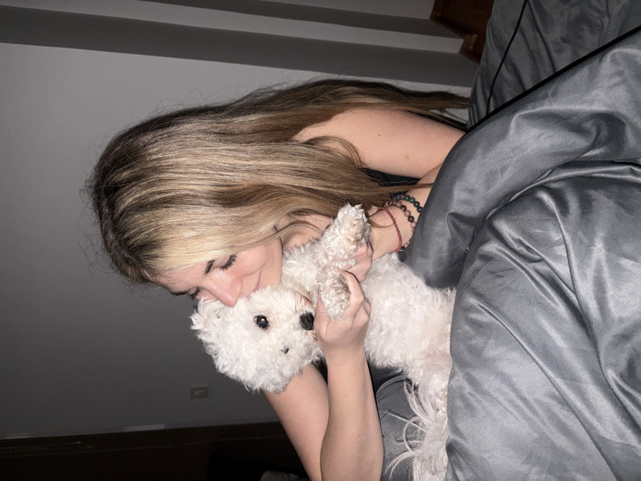 A woman with long blonde hair embraces a small, fluffy white dog. She is lying in bed under gray bedding. The woman wears several bracelets on her right wrist. The dog's face is buried in her neck. The scene is lit dimly.