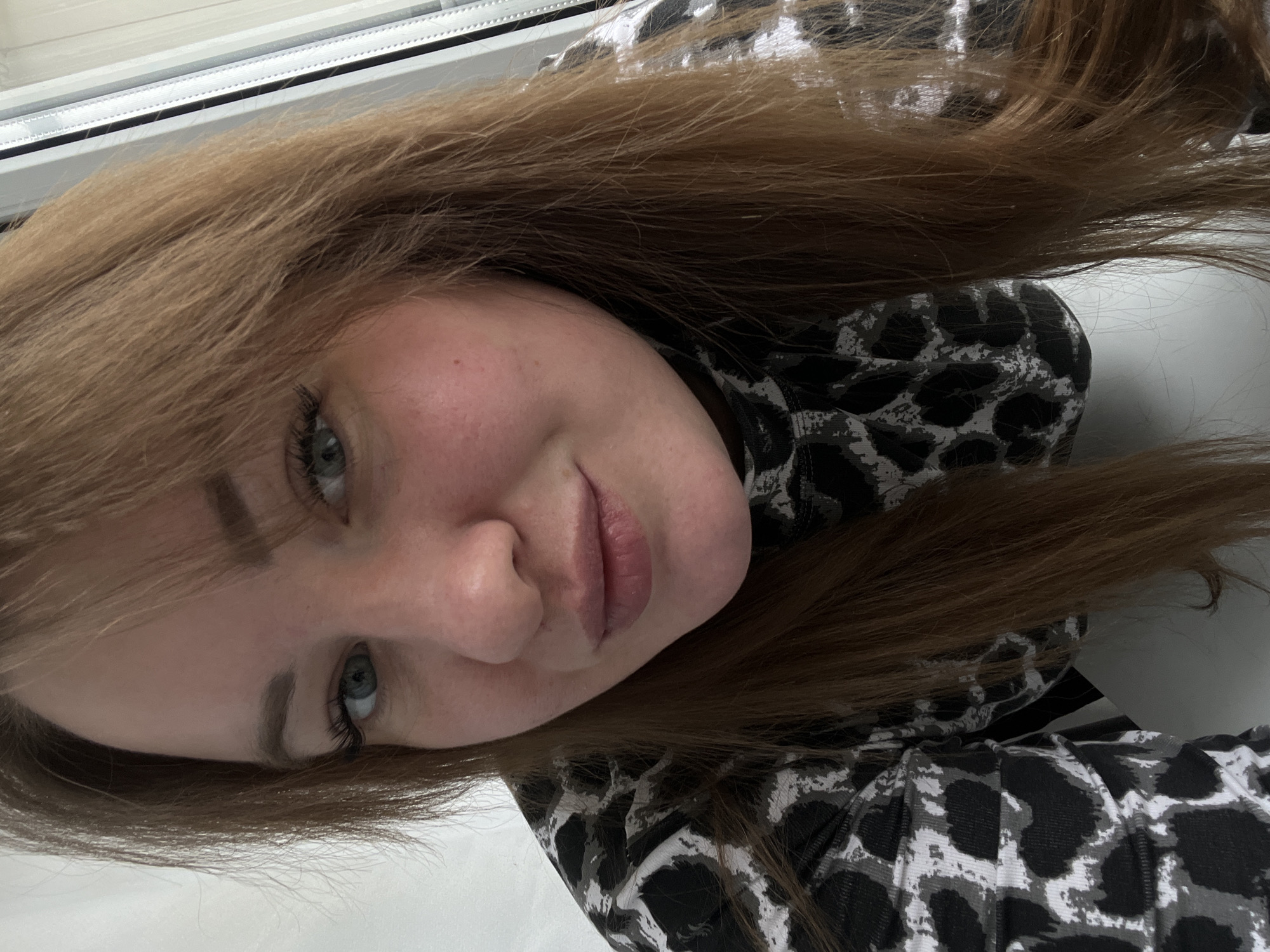 A young woman with long brown hair is shown in a close-up selfie. She has fair skin, blue eyes, and pink lips. She is wearing a gray and black patterned top. The background includes a white windowsill. The location is not evident from the image.