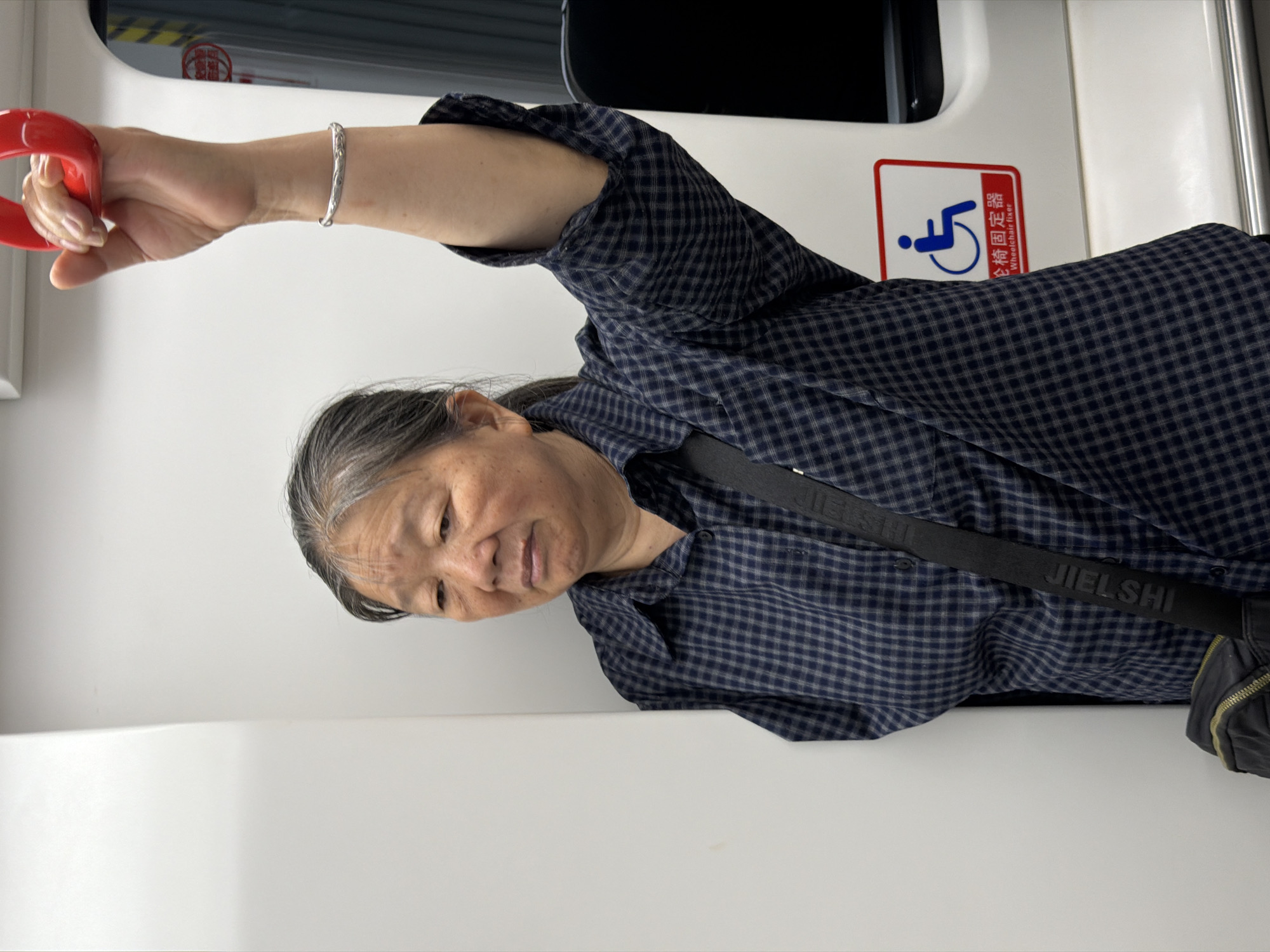 An elderly East Asian woman stands inside a modern subway car, identified by the presence of overhead red handles and a specific accessibility sign. Her left side is pressed against a flat, white interior panel of the train. The woman has grey hair, visible wrinkles on her face and forehead, and a neutral expression with a slight downward gaze. She is wearing a dark blue and light blue/grey plaid long-sleeved button-down shirt; the sleeve on her right arm is partially rolled up. A silver-colored bangle adorns her right wrist. Her right arm is raised, and her hand grasps a red, ergonomic overhead strap. A black cross-body bag with a visible strap bearing the white text "JIELSHI" and a dark zipper crosses her torso. To her right, on the white interior panel, is a rectangular red and white sticker featuring a blue wheelchair icon and Chinese text "轮椅固定器" (Lúnyǐ gùdìngqì) with the English translation "Wheelchair fixer" below it. A dark vertical strip, possibly a window or door frame with yellow and black safety markings, is visible further to the right. Based on the Chinese signage, the scene is located in China, specifically within the context of a public transport system in the Yuzhong District.