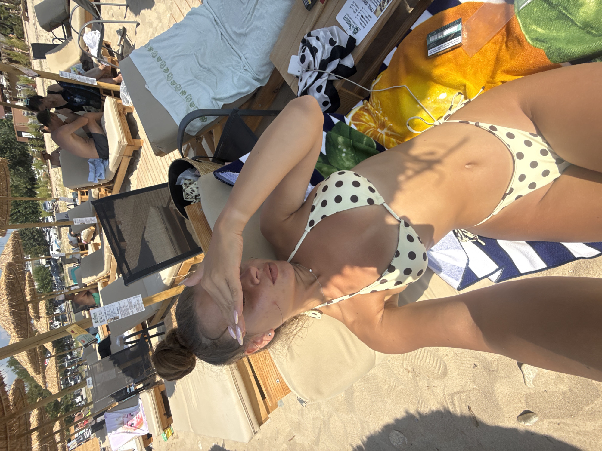 A young woman is lying on a beach chair on Ofrynio Beach, Greece. She is wearing a polka dot bikini and has her hand covering her eyes. The beach is lined with umbrellas and other beach chairs. There are other people in the background, some of whom are also sitting on beach chairs. The water is not visible in the frame. The sand is light brown and there are a few small rocks visible. The woman's hair is pulled up into a bun.