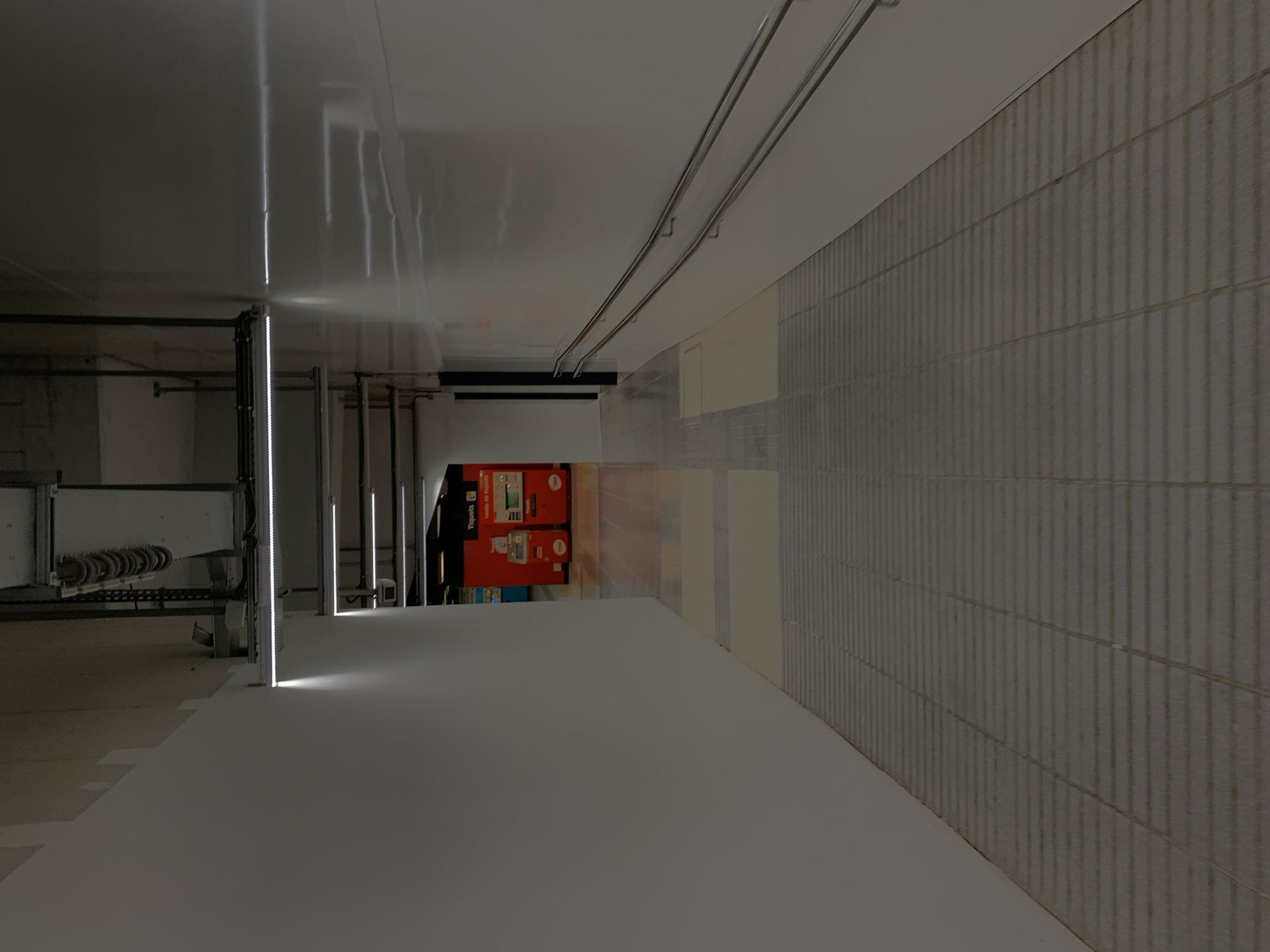 The image depicts an underground section of a metro station in Barcelona, Spain. The scene is illuminated by artificial lighting, with a focus on the clean, gray-toned aesthetics. A metal beam and ductwork run along the ceiling, complemented by a tiled wall on the right-hand side. On the left, there is a visible train track area, with metal structures and some machinery. In the background, a ticket vending machine stands in an adjacent space. The area is empty of people, emphasizing the cold, minimalist architecture of the station.