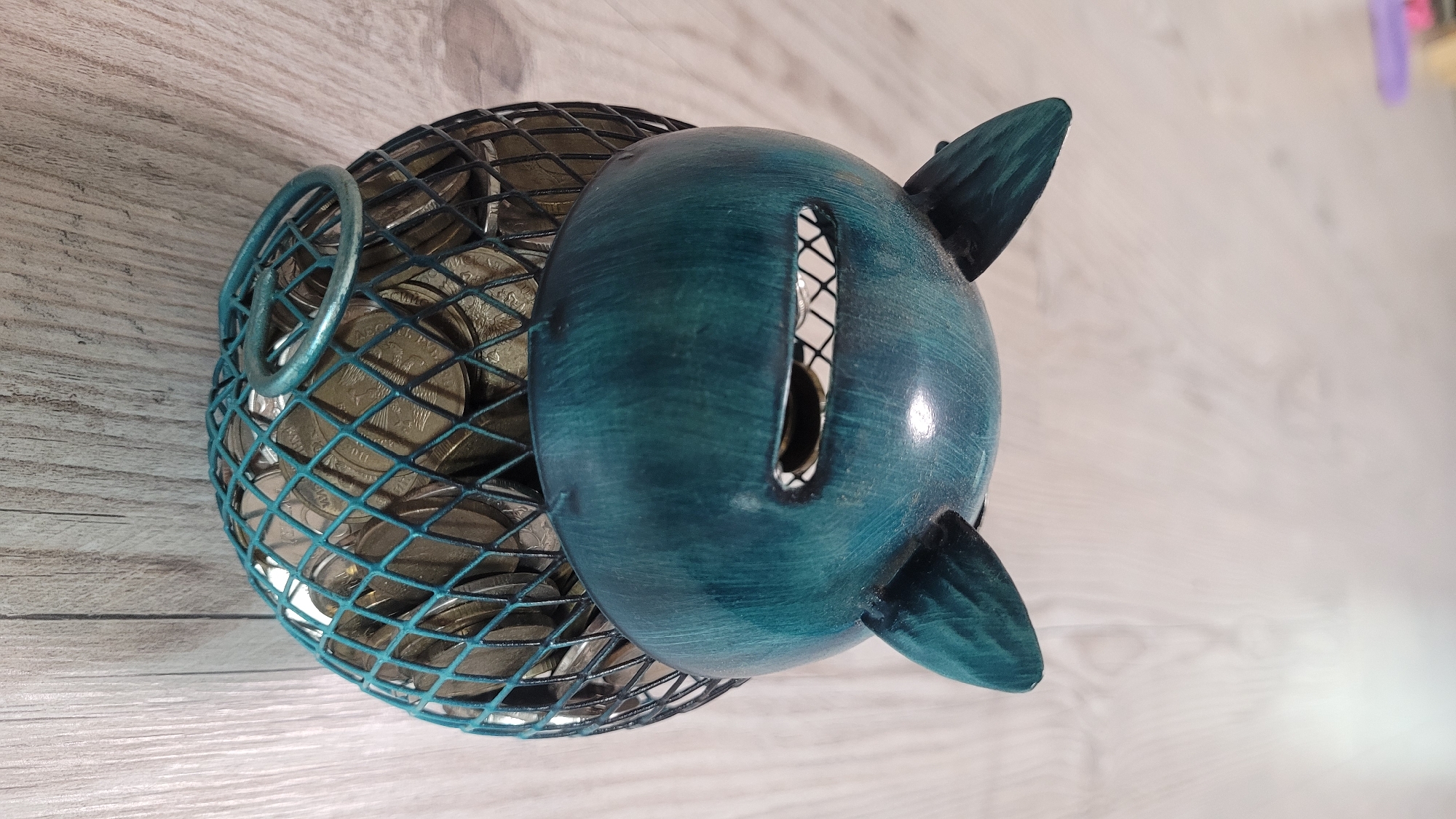 A teal-colored, animal-shaped piggy bank, resembling a cat or owl, sits on a light-colored wooden floor. The piggy bank has two pointed ears, a rectangular slot for coins with a jagged edge resembling teeth, and a wire-mesh body that reveals a collection of coins inside. A small, circular handle is attached to the front of the wire-mesh body. The background is blurred, with hints of purple and a light source creating a bright area. The scene is static and appears to be indoors, in an unspecified urban location.