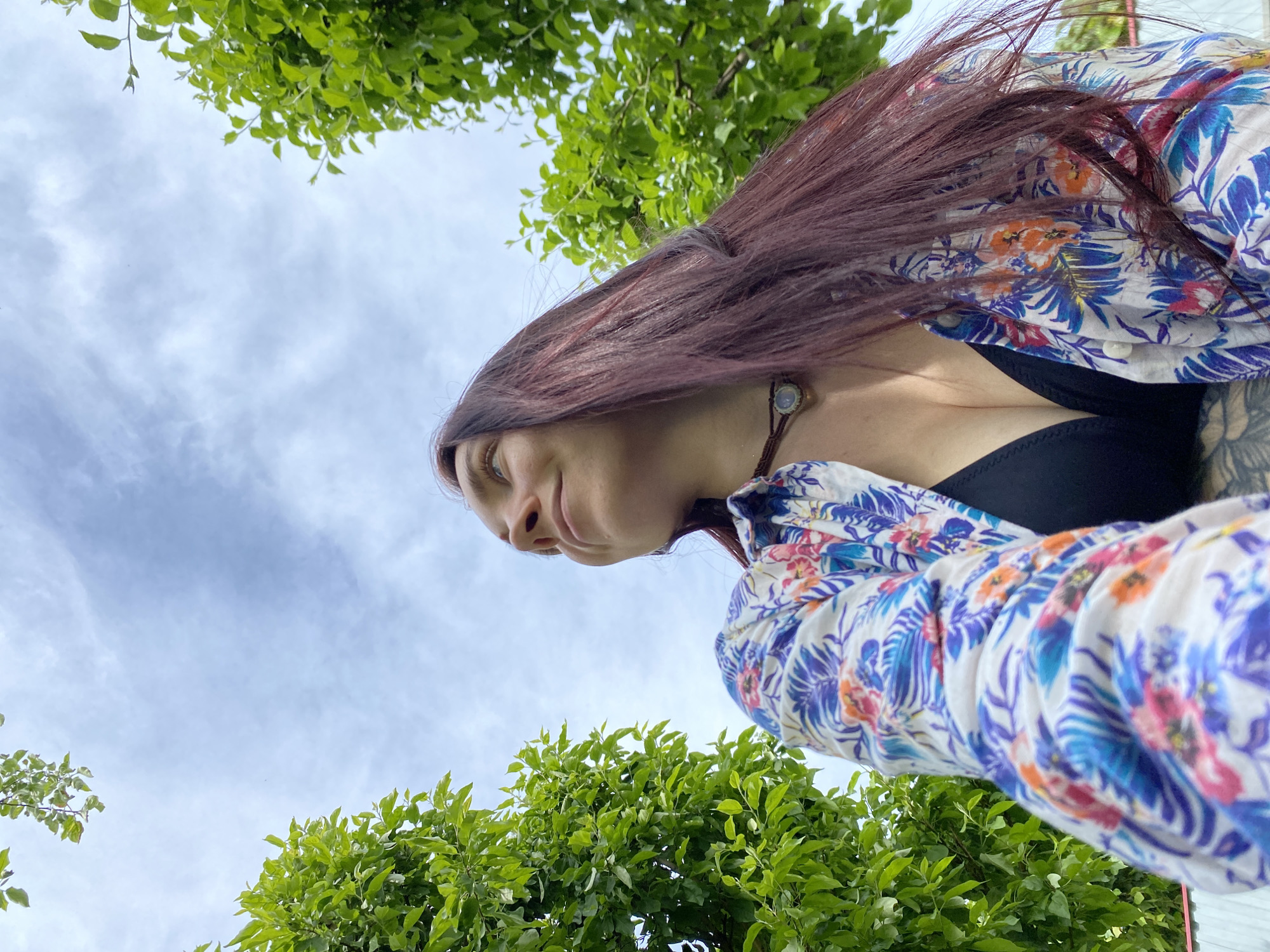 The image presents a low-angle outdoor portrait of a woman with long, dark reddish-brown hair. She is positioned in the right half of the frame, looking slightly to her left (our right) with a subtle, calm expression. She is wearing a loose-fitting, button-up shirt with a vibrant tropical floral pattern, featuring blue, purple, orange, and pink foliage and flowers on a white background. Beneath the open shirt, a black top or bra is visible. A circular pendant on a brown cord hangs around her neck, and part of a tattoo is visible on her right upper arm or shoulder.
The perspective is directed upwards, with the woman framed against a bright, cloudy sky that occupies the central background. Lush green foliage, likely from trees or bushes, borders the top edge and the lower left corner of the image, suggesting a natural outdoor setting such as a park or garden. The scene appears to be taking place during the daytime, under an overcast sky. No text is visible in the image. This scene is located in Chaikovsky, Russia.