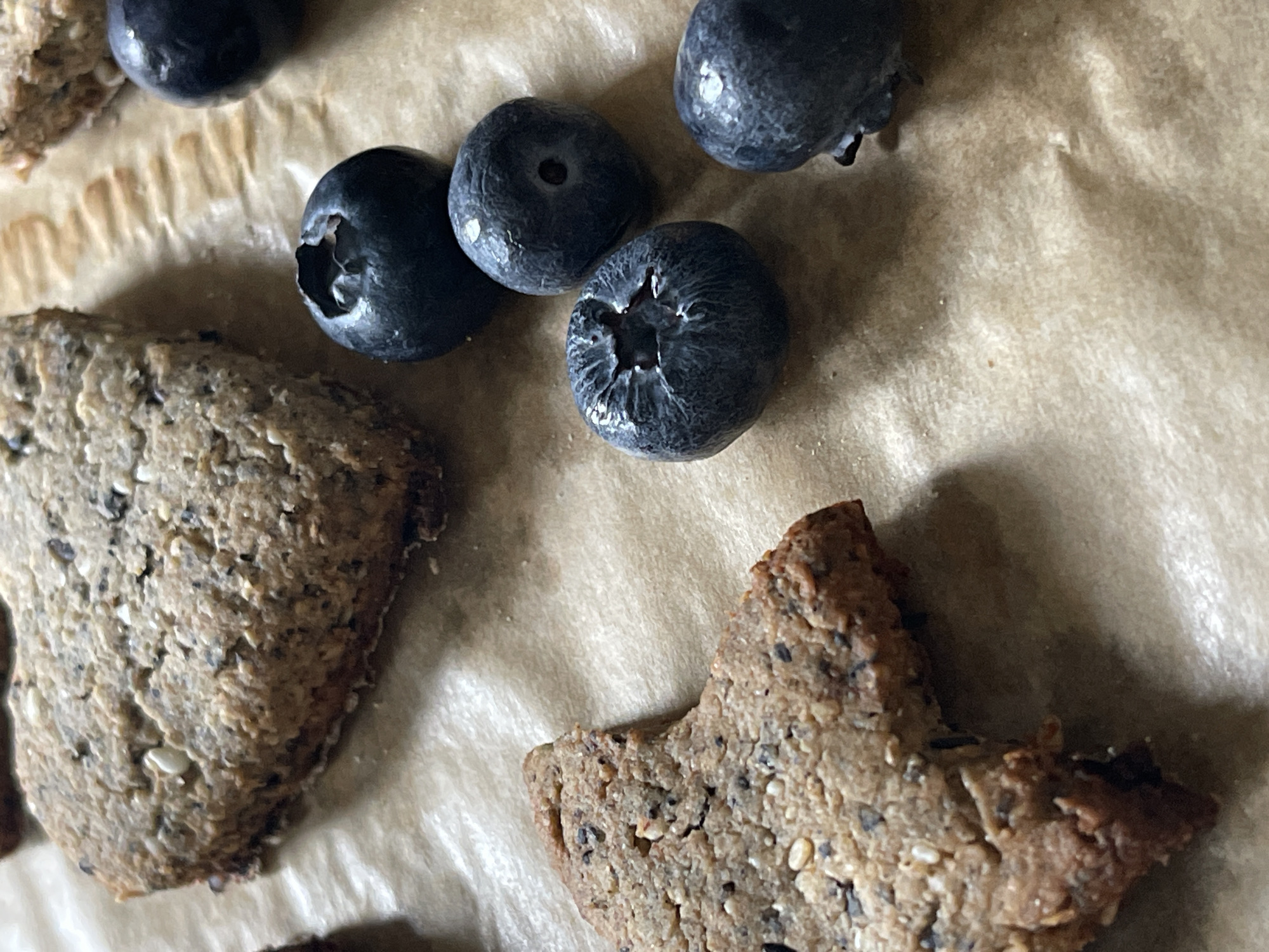 The image shows a close-up of blueberries and two baked goods on a piece of parchment paper. The blueberries are scattered amongst the baked goods. The baked goods appear to be cookies or small cakes. The background is blurred.