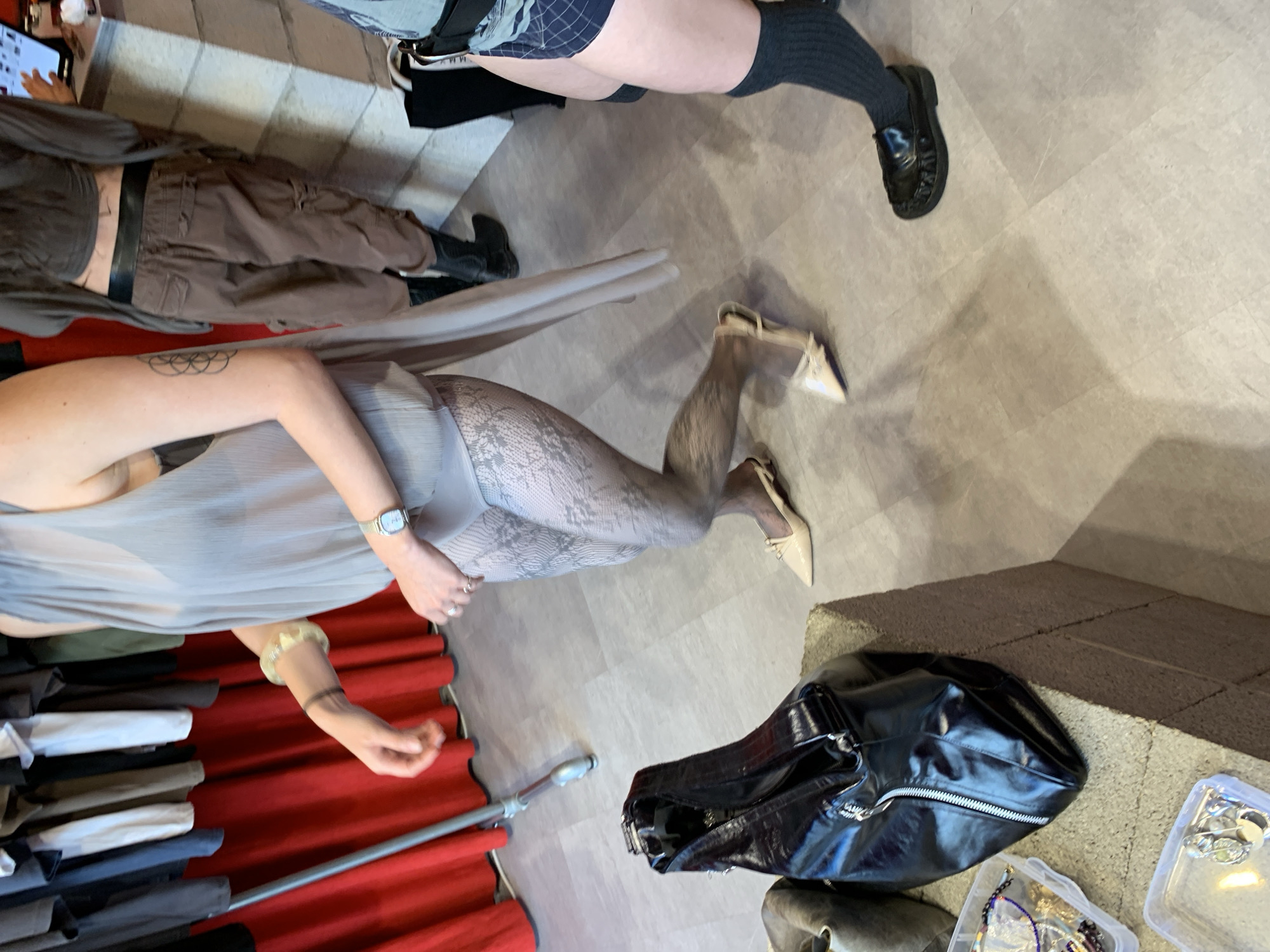 This image captures an indoor scene, likely a retail space or studio in Tel-Aviv, Israel, presented rotated clockwise. The perspective is from above, looking down on several partially visible individuals and various items.
In the lower-left section of the frame, a person is featured, wearing a sheer, light grey pleated top or dress over a dark bra. They are adorned with light grey lace-patterned tights and light-colored pointed heels featuring small bows. A silver watch is visible on their left wrist, a light-colored bracelet on their right, and a geometric tattoo on their upper right arm. Behind this individual, a clothing rack with red fabric lining holds stacks of folded garments in various neutral shades like white, black, grey, and brown.
Towards the top-left, another person is partially seen, wearing a dark top, dark brown cargo pants with pockets, and black boots. Their midriff is exposed, revealing a tattoo on their left upper arm. A hand holding a phone or tablet displaying text or code is also visible in this area. In the top-right, the lower body of a third person is shown, dressed in blue and white plaid shorts, dark ribbed socks, and shiny black loafers.
On the floor in the lower-right corner, a glossy black bag with a silver zipper rests next to two clear plastic containers filled with an assortment of jewelry, including necklaces and rings. The floor is a light grey, tiled or patterned surface, while a darker, rough-textured concrete or stone structure is partially visible on the far right. The overall activity appears to be casual, possibly browsing or styling in a shop or studio setting during the daytime. No distinct text is clearly legible, aside from faint details on the phone screen and the black bag.