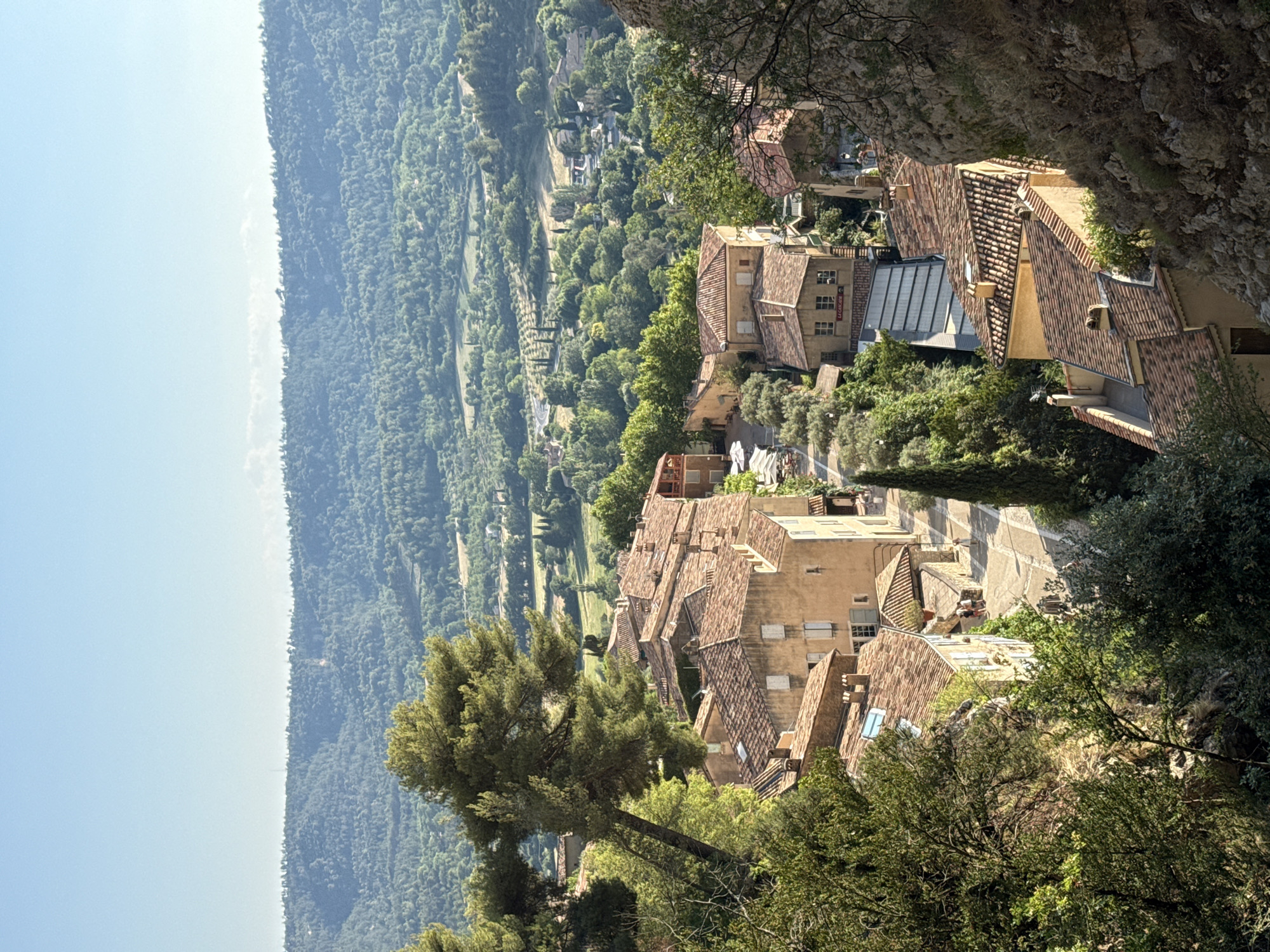 This is a high-angle, medium shot of a village nestled in a valley, with rolling hills and lush green trees in the background. The village features buildings with terracotta tile roofs and light-colored walls, characteristic of Moustiers-Sainte-Marie, France. In the foreground, a large pine tree leans into the frame from the left, with its branches obscuring some of the village's rooftops. The buildings are densely packed, with narrow streets winding between them. Some buildings have awnings or outdoor seating areas, suggesting commercial establishments. A few people are visible walking on the streets. The sky is clear and blue, indicating a sunny day. The overall impression is of a charming, sun-drenched village in a picturesque natural setting.