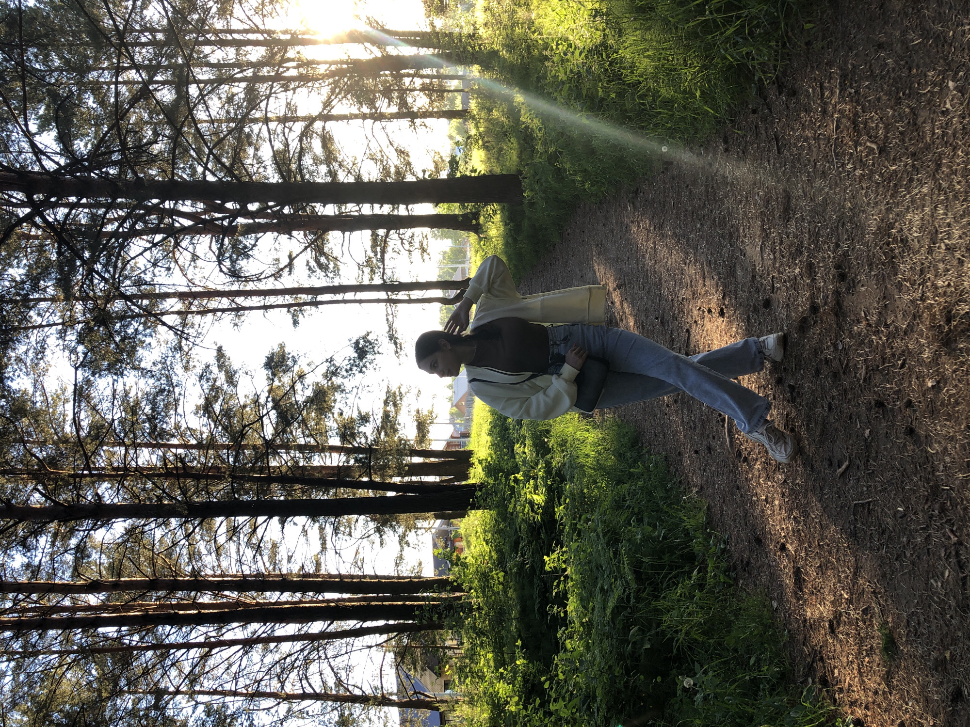 A young woman is walking on a dirt path through a forest. The path is lined with trees on both sides, and the sun is shining through the trees, creating dappled light on the ground. The woman is wearing a black shirt, blue jeans, a white jacket, and white shoes. She is looking to the left and is touching her hair with her right hand. The location may be the city of Chaikovsky, Russia, based on the query.