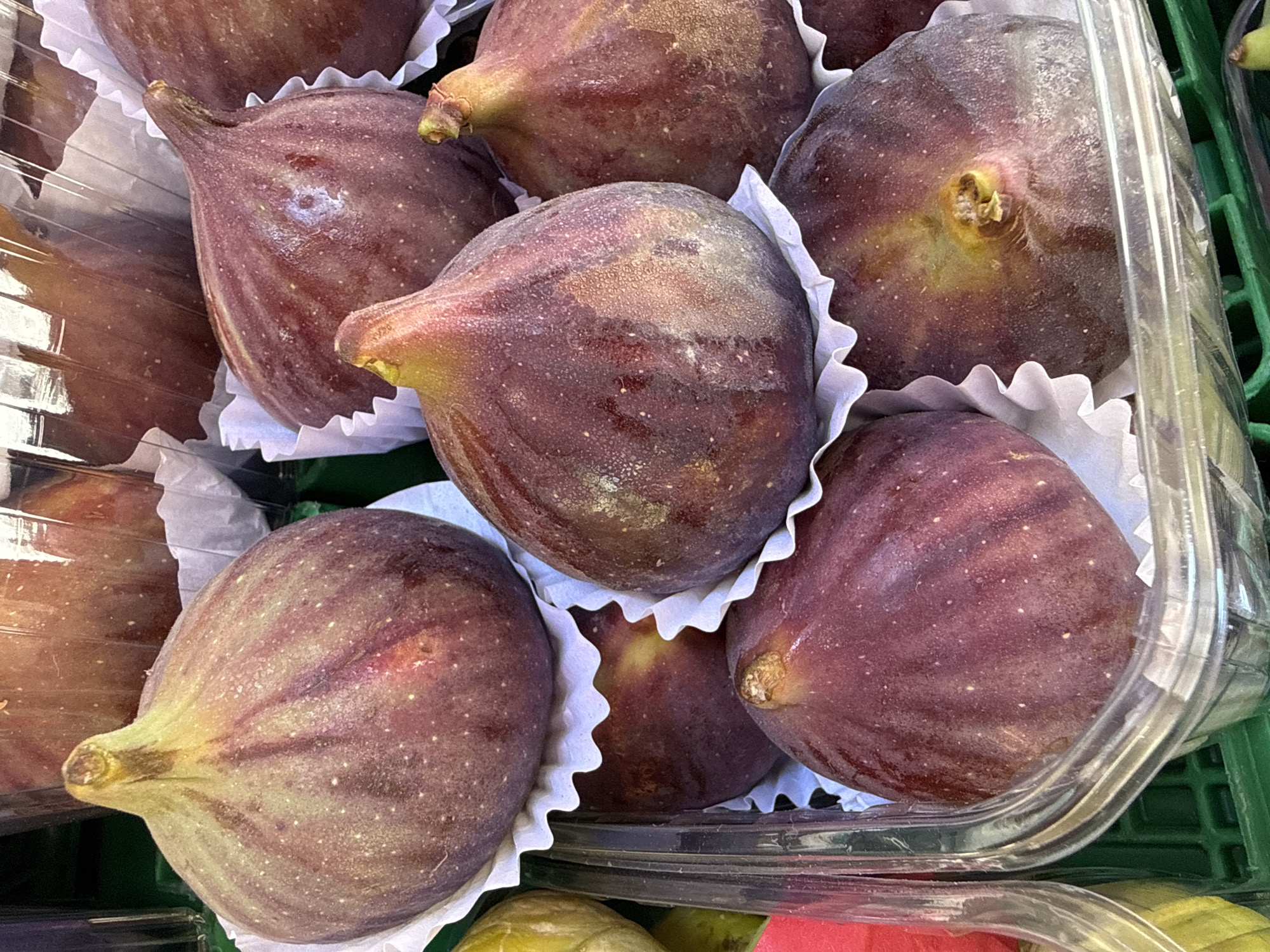 The image shows a close-up of ripe figs displayed in clear plastic containers, resting on a green market stall. The figs are a deep purple-red color with visible white speckles and a slightly dry, powdery bloom. Each fig is nestled in white paper liners, which are ruffled around the edges. In the background, more figs are visible, some in similar packaging, and a hint of a larger market environment. The lighting is bright, highlighting the texture and sheen of the fruit. The scene is likely from a street market or produce stand in Kadıköy, a district known for its vibrant markets and a popular spot for fresh produce in Istanbul, Turkey.