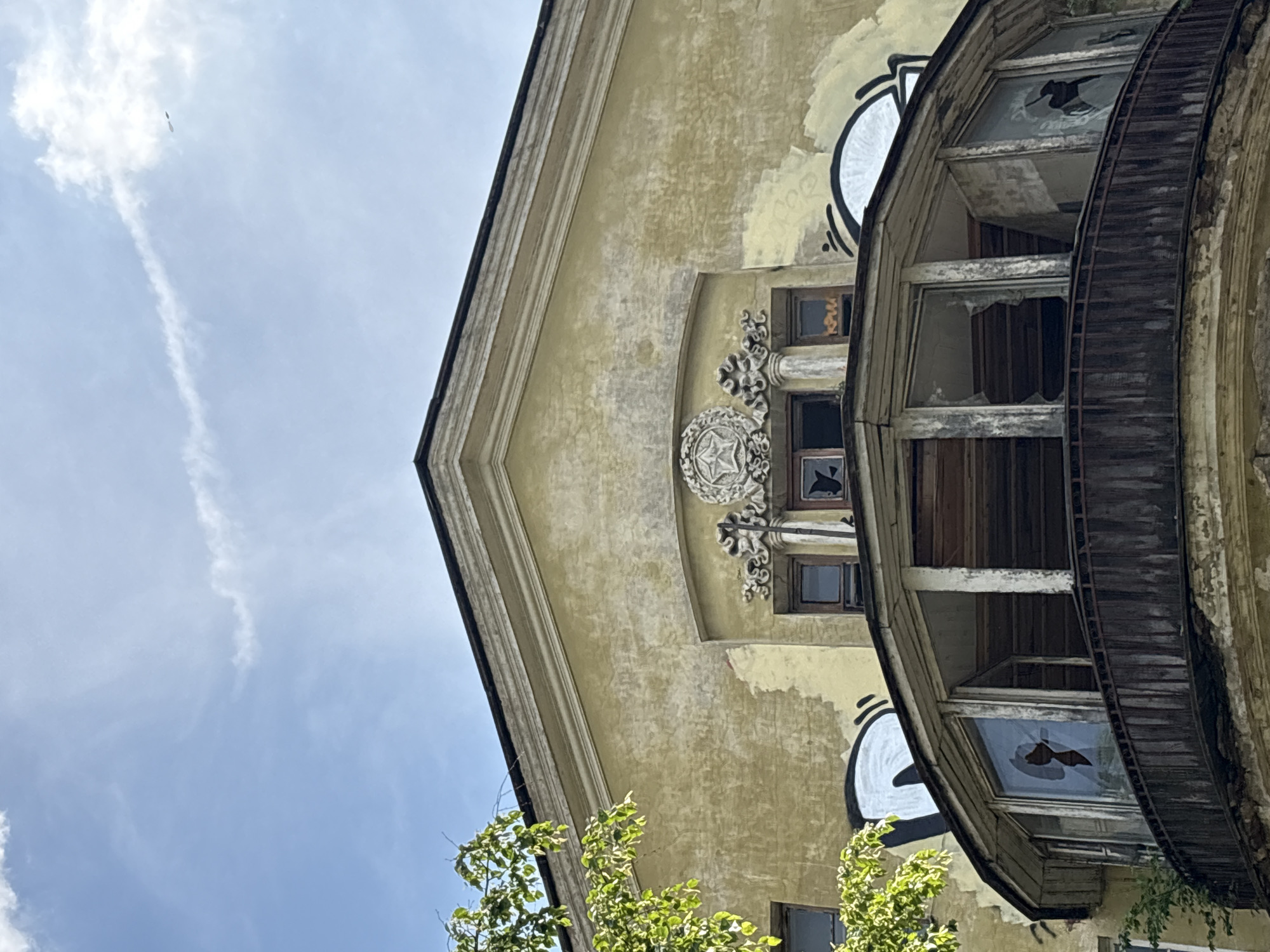 The image shows the upper facade of a deteriorating building with a prominent semicircular balcony. The building's yellow plaster is peeling and stained, with graffiti visible on the upper walls, including large white and black abstract shapes and what appears to be Cyrillic lettering in orange. The balcony features broken glass panes and a weathered wooden railing. Above the balcony, there are several windows, some with cracked glass, and ornate architectural details, including a star emblem. A tree with green foliage partially obscures the left side of the building. The sky above is blue with scattered clouds and a faint contrail. The overall impression is one of neglect and urban decay. This specific building is located at Liteyny Prospekt 55 in Saint Petersburg, Russia, and is known for its dilapidated state and distinctive architectural features.