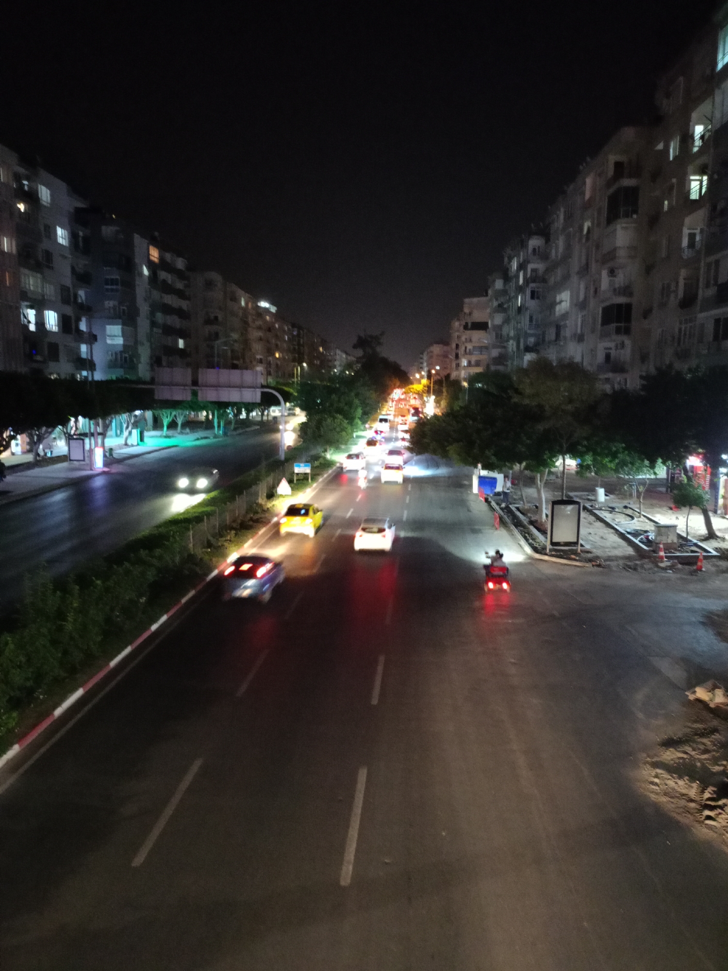 A nighttime, high-angle shot captures a street scene in Antalya, Turkey, likely on a main thoroughfare given the multi-lane road and apartment buildings lining both sides. The road is wet, reflecting the light from streetlights and vehicle headlights, creating streaks of illumination.
Several vehicles are in motion, their lights blurred due to the long exposure. A yellow taxi is visible in the right lane, and a blue car with red taillights is in the left lane. Further down the road, other cars and a red-lit motorcycle or scooter are present. The buildings on either side are residential apartments, with some windows illuminated, suggesting occupancy. Trees line the sidewalk on the left side of the road, and a grassy median separates the lanes in some areas. Construction materials and traffic cones are visible on the right side of the road. The sky is dark and clear.