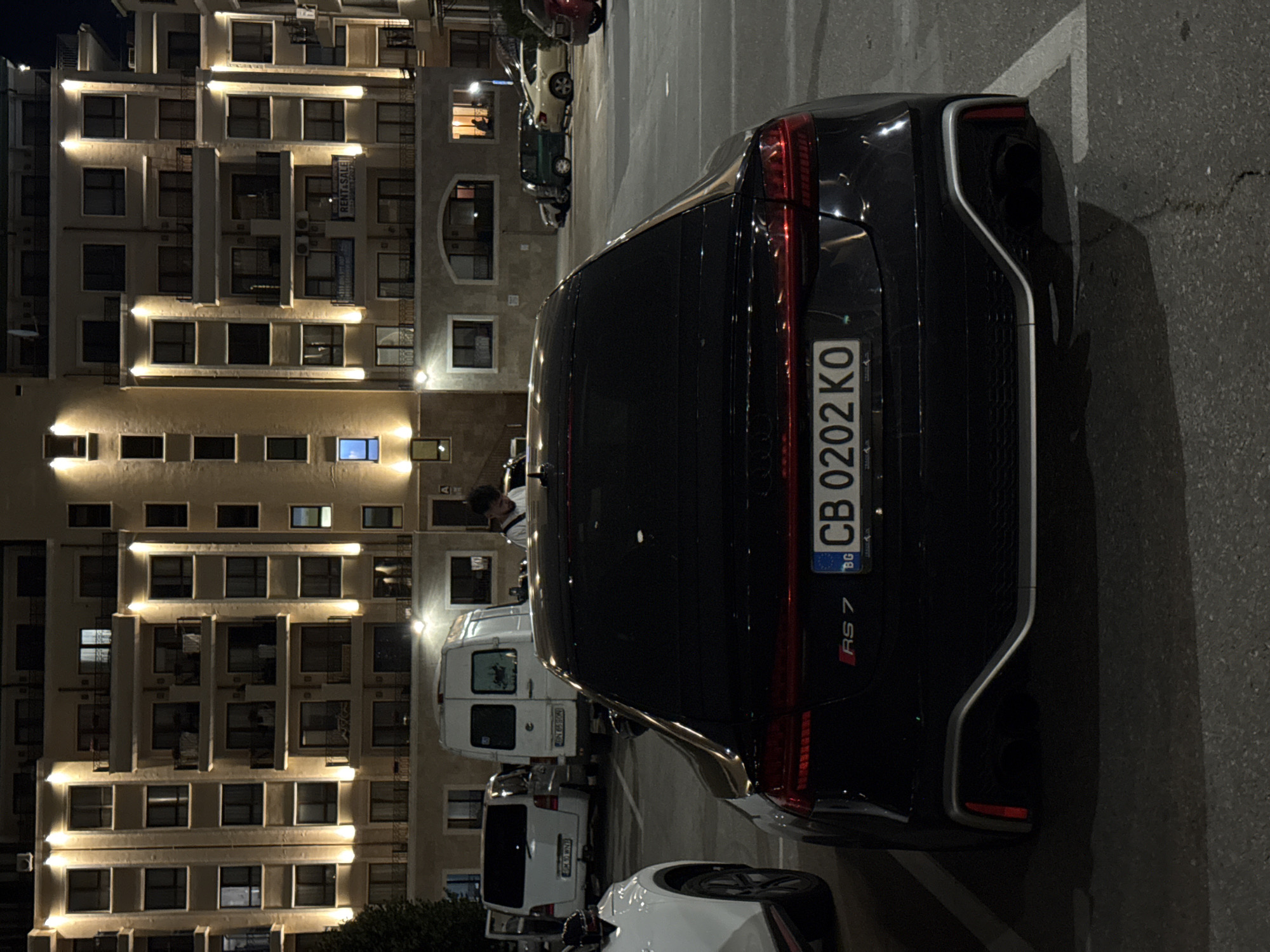A black Audi RS 7 is parked in a paved lot at night. The car faces away from the camera, and its license plate reads "CB 0202 KO". Behind the Audi, several other vehicles are parked, including a white van. In the background, a multi-story building is illuminated by external lights. One person is visible on the second floor of the building, looking out a window. Another person, a young male with dark hair, stands near the white van. The environment appears to be an urban setting, likely a residential or commercial area in Varna, Bulgaria. The lighting suggests it is after dark.