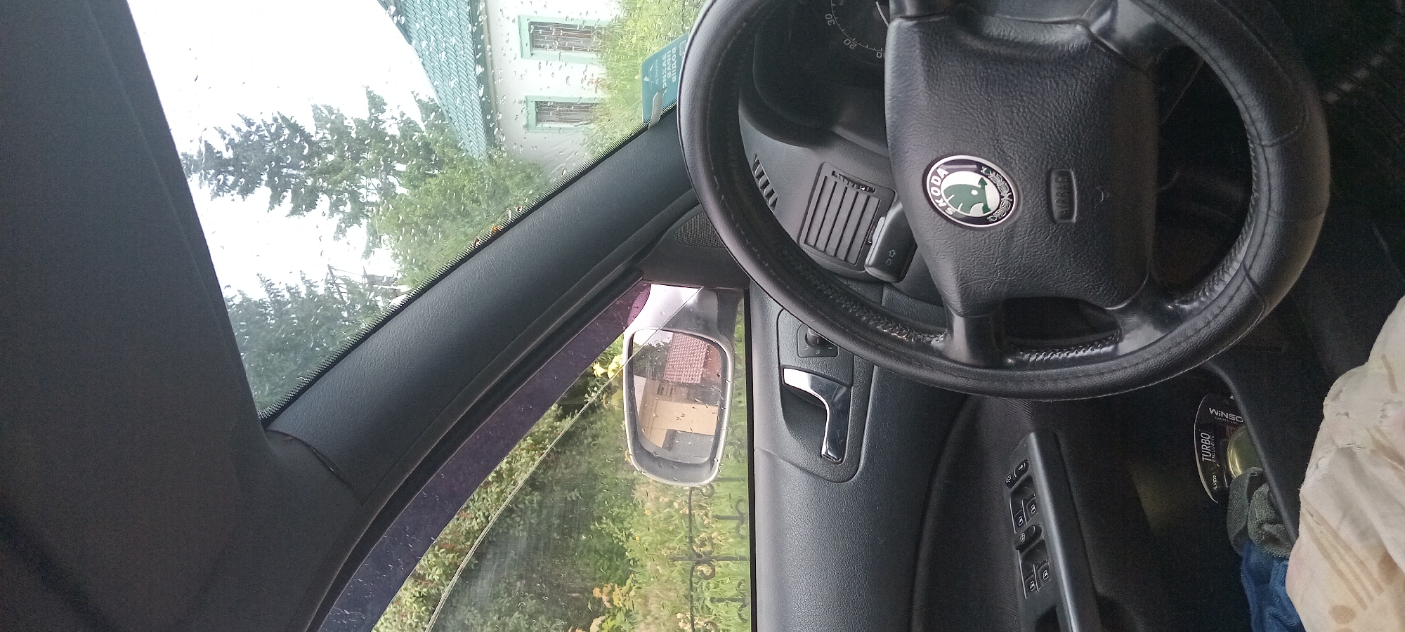 The image shows the interior of a car, looking out the driver's side window. The steering wheel, dashboard, and door panel are visible. Outside, there is a house with a green roof, trees, and greenery. The window has raindrops on it, suggesting it might be raining. A side mirror reflects a part of the house. In the foreground, there are some items in the car's storage compartment, including a package labeled "TURBO EXCLUSIVE." The scene appears to be in a residential area, possibly on the outskirts of Koviahy, Ukraine, given the context provided. There are no people visible in the image. The overall impression is of a quiet, possibly overcast day.