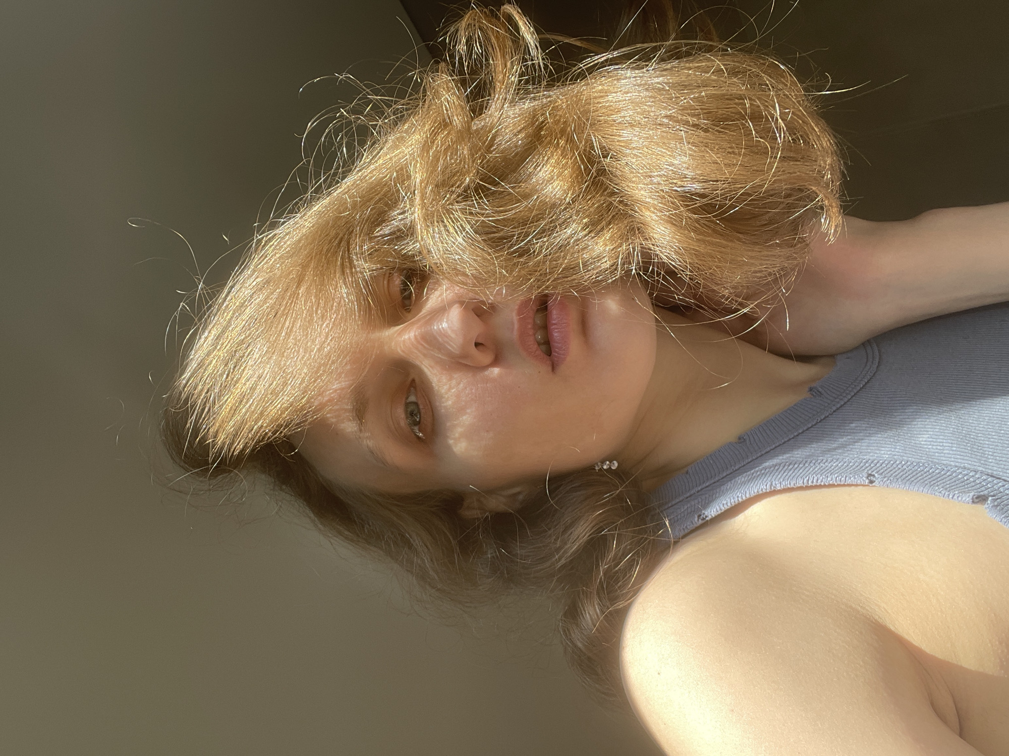 A woman with reddish-blonde hair is positioned with her head tilted back and to the side, her face slightly obscured by her hair. She wears a light blue sleeveless shirt. Part of her right shoulder and upper chest are visible. A hand is touching her neck. The background is a light gray color. The lighting is strong and uneven, creating distinct shadows on her face and neck. The location is not identifiable as Подшивалово, Russia, from the visual information provided.