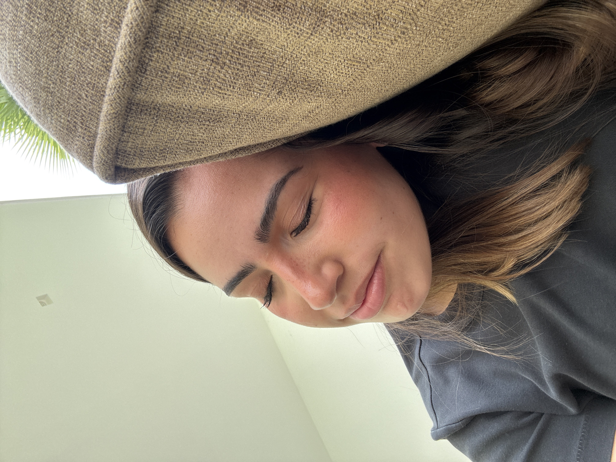 A young woman with light skin, brown hair, and closed eyes is positioned under a brown upholstered object, possibly a piece of furniture. She is wearing a dark-colored shirt. The background shows a light-colored wall. The location is implied to be in San Bernardino Tlaxcalancingo, Mexico.