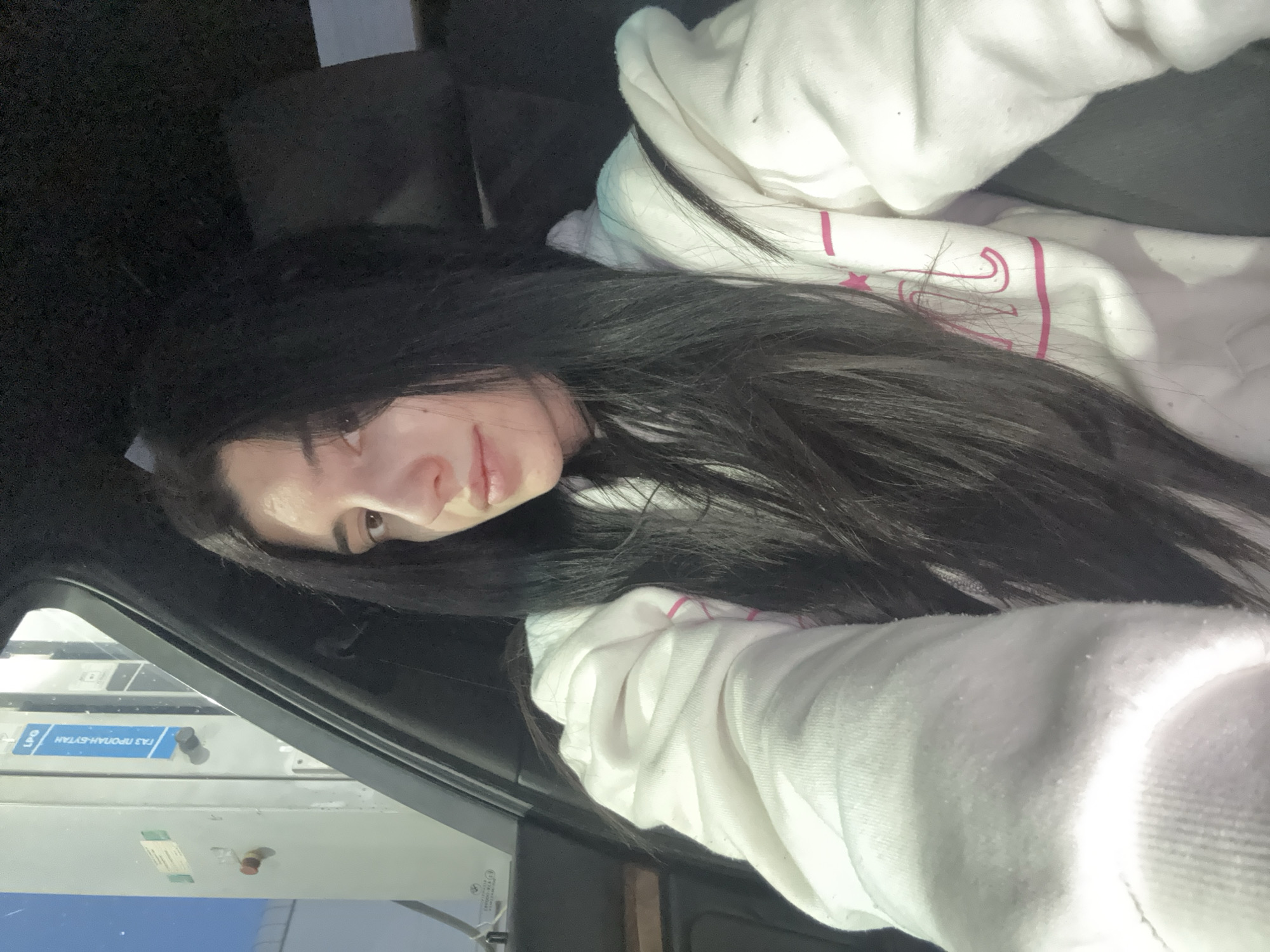 A young woman with long, dark hair is reclining inside a vehicle. She is wearing a white hoodie. Part of the vehicle's interior is visible, including the dashboard and a window. Outside the window, there's a view of what seems to be a gas pump or utility station, identifiable by its blue signage and metallic structure. It reads, "ГАЗ ПРОПАН-БУТАН" in Cyrillic, which translates to "LPG PROPANE-BUTANE". Given the Cyrillic text, the location is likely in Bulgaria. Since the query requests a location in Sofia, Bulgaria, it suggests the photo was taken at a gas station in Sofia that sells LPG. However, the exact street address and neighborhood cannot be ascertained from this image alone.