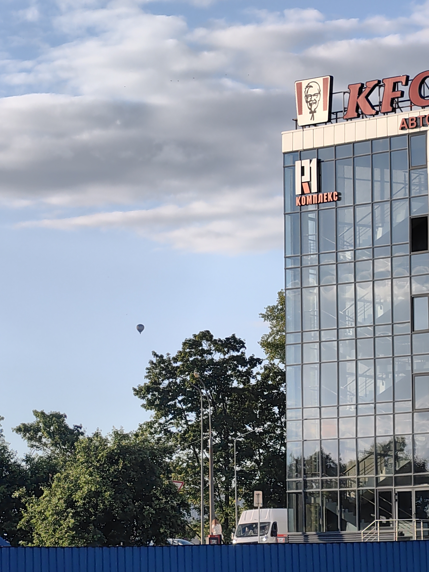 The image captures a sunny day in the city of Fanipolski Sel'ski Savet, Belarus. Dominating the right side of the frame is a modern glass-fronted building, likely a commercial establishment, with the prominent signage of KFC visible at the top, featuring the Colonel Sanders logo. Below the KFC sign, the word "АВТО" (AUTO) is also visible. A smaller sign on the building's facade reads "Р| КОМПЛЕКС" (R| COMPLEX). The building's reflective windows show the sky and trees. To the left, a bright blue fence runs along the bottom of the image. In the sky, a hot air balloon is visible in the distance. Several trees, one with a lamppost in front of it, are also present on the left and right sides of the scene. A white van is parked in front of the building. A person is standing near the van, and another individual is walking in the background. The sky is blue with scattered clouds.