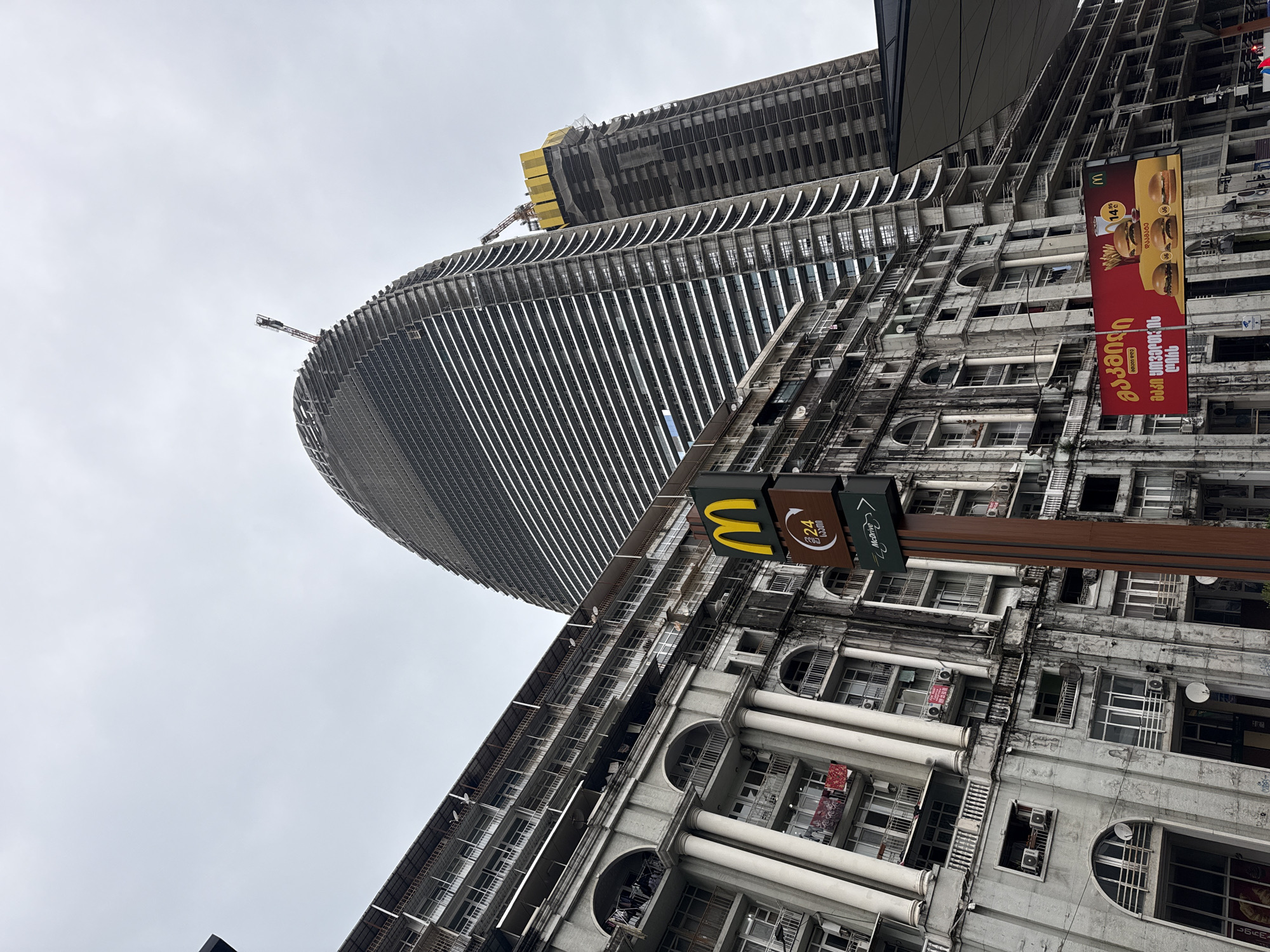 The image captures a unique juxtaposition of architectural styles in the city of Batumi, Georgia. In the foreground, a weathered, Soviet-era apartment building dominates, characterized by its repetitive arched windows, white columns, and aging concrete facade. A McDonald's sign is affixed near the bottom. Behind and towering over the apartment complex is a modern skyscraper under construction. The high-rise's curved form and horizontal lines create a striking contrast to the older structure. The sky is overcast and grey.
