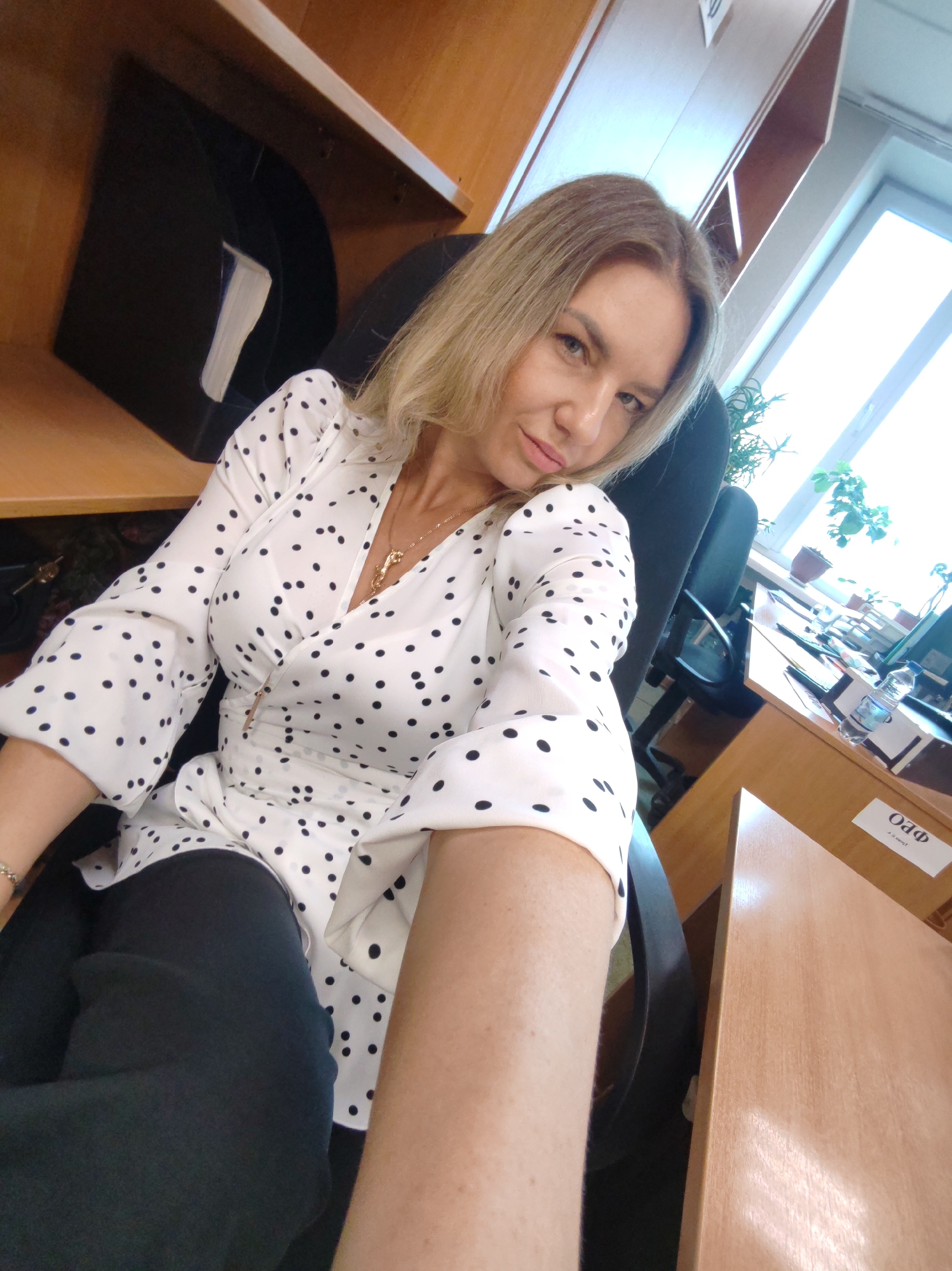 A woman is captured in a selfie taken within an office environment in Petropavlovsk-Kamchatsky, Russia. She has blonde hair, fair skin, and is looking directly at the camera with a neutral to slight smiling expression. She is wearing a white blouse adorned with black polka dots, a gold-colored necklace, and dark trousers. Her arm is extended, indicating she is holding the camera for the self-portrait.
She is seated in a black office chair, positioned next to a wooden desk. The setting features light wooden office furniture. To her left, a black filing tray holding white papers is visible on a shelf. To her right, a large window allows bright daylight into the room, illuminating several potted plants on the windowsill. A computer monitor and a bottle of water are also present on the desk surface.
A small white sign is affixed to the side of the desk, clearly displaying the Cyrillic text "ОФО" in large letters. Below it, smaller text is visible, which appears to read "Что это такое?" (What is this?). The bright natural light streaming through the window suggests it is daytime. The image portrays a moment of casual interaction within a professional workspace.