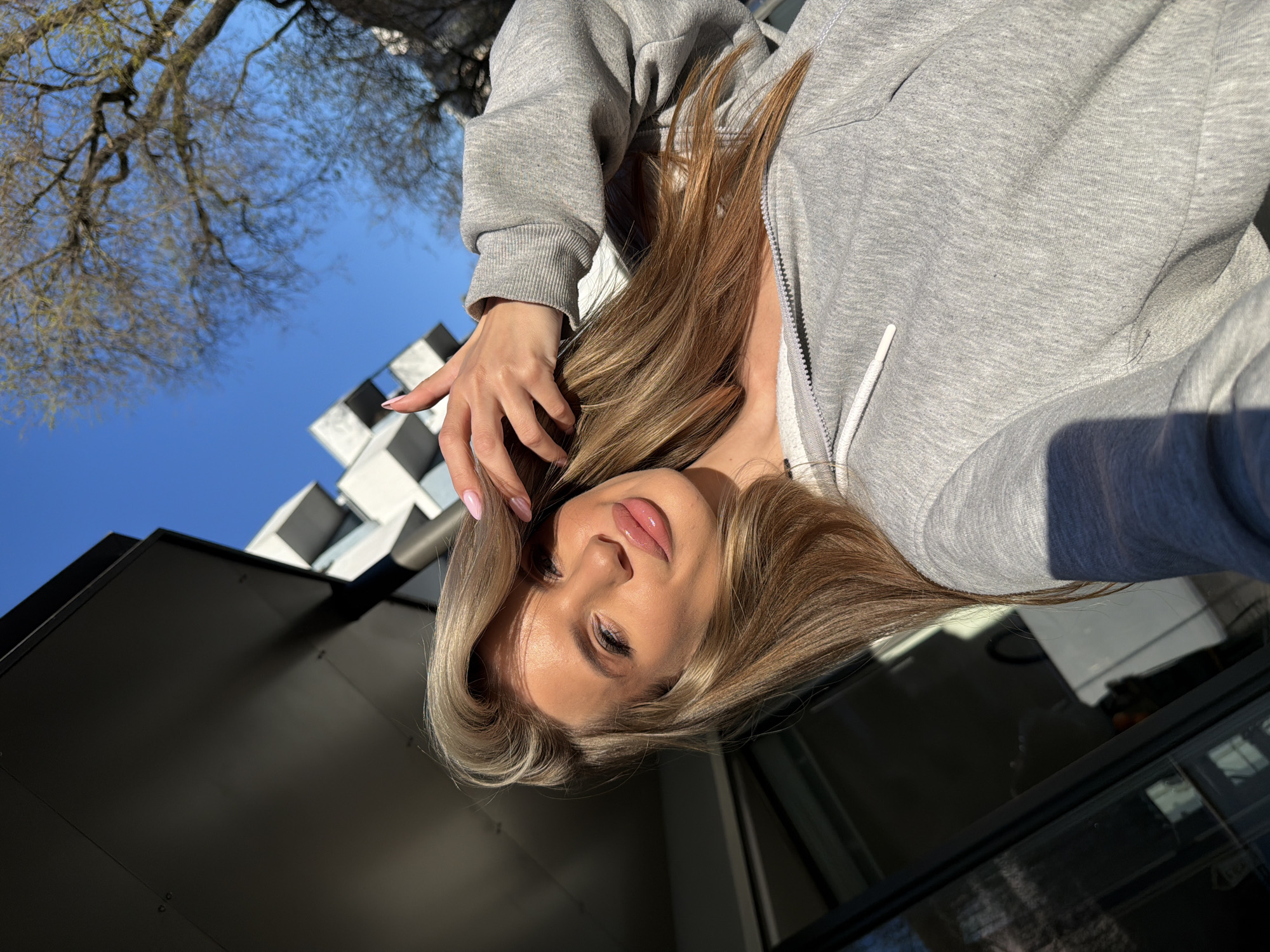 A selfie is taken from a low-angle perspective with a fair-skinned, blond-haired woman wearing a gray hooded sweatshirt as the primary subject. She is looking directly at the camera with her hand near her face. The backdrop includes a clear blue sky and architectural features of Berlin. The image suggests an urban location, possibly a city street or plaza. The presence of a modern building design in the background indicates a contemporary part of Berlin. A tree with leafless branches can be seen in the upper left corner.