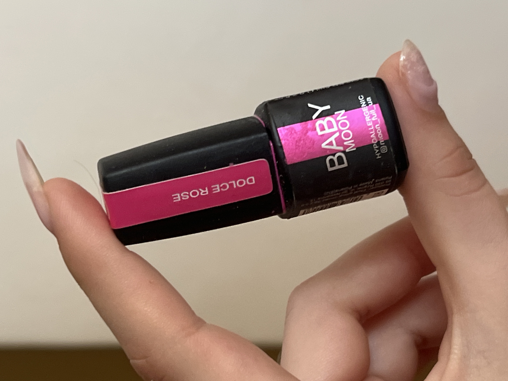 A person's hand is holding a small black bottle of nail polish. The bottle has a pink label with white text that reads "BABY MOON" and "DOLCE ROSE". Below that, it says "HYPOALLERGENIC" and an Instagram handle "@moon_full_ua". The person's fingernails are visible and appear to be manicured. The background is a light-colored, out-of-focus surface, possibly a wall. The lighting suggests the image was taken indoors, possibly in a domestic setting. There are no discernible landmarks or environmental cues to suggest a specific city or location.