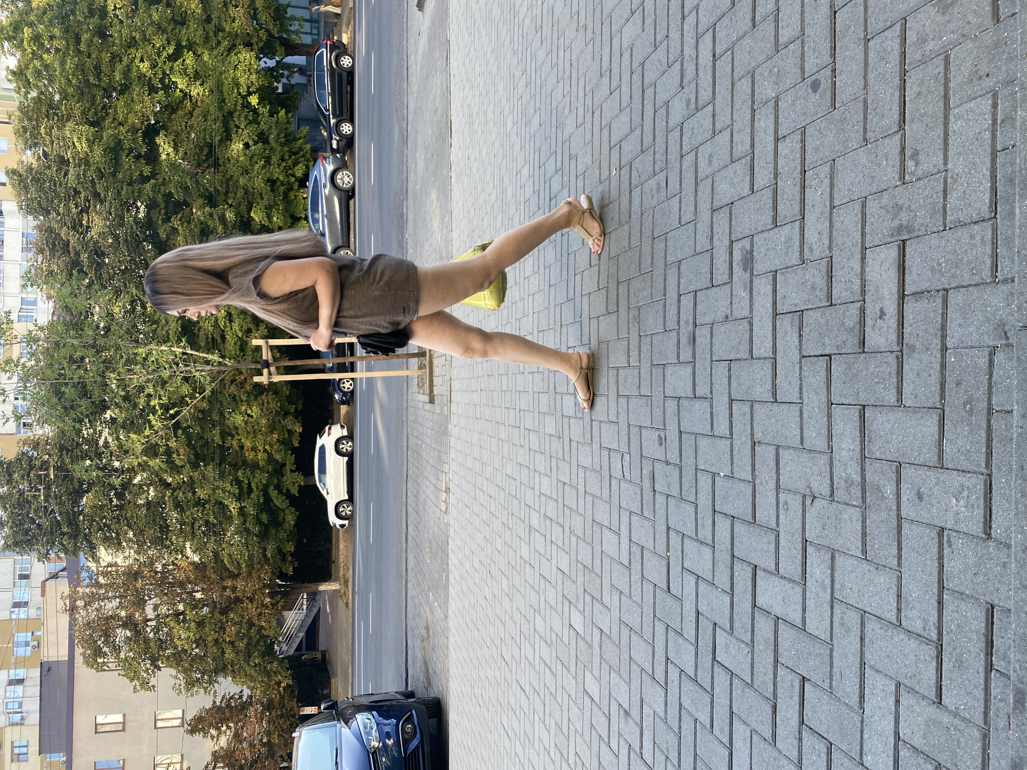 A woman with long, blonde hair walks on a paved sidewalk in Chișinău, Moldova. She is wearing a brown sleeveless dress and light brown sandals. A yellow bag is slung over her right shoulder, and she carries a black purse on her left. In the background, trees line the street, with cars parked along the roadside and buildings visible beyond. The scene appears to be a typical urban street during daylight hours.