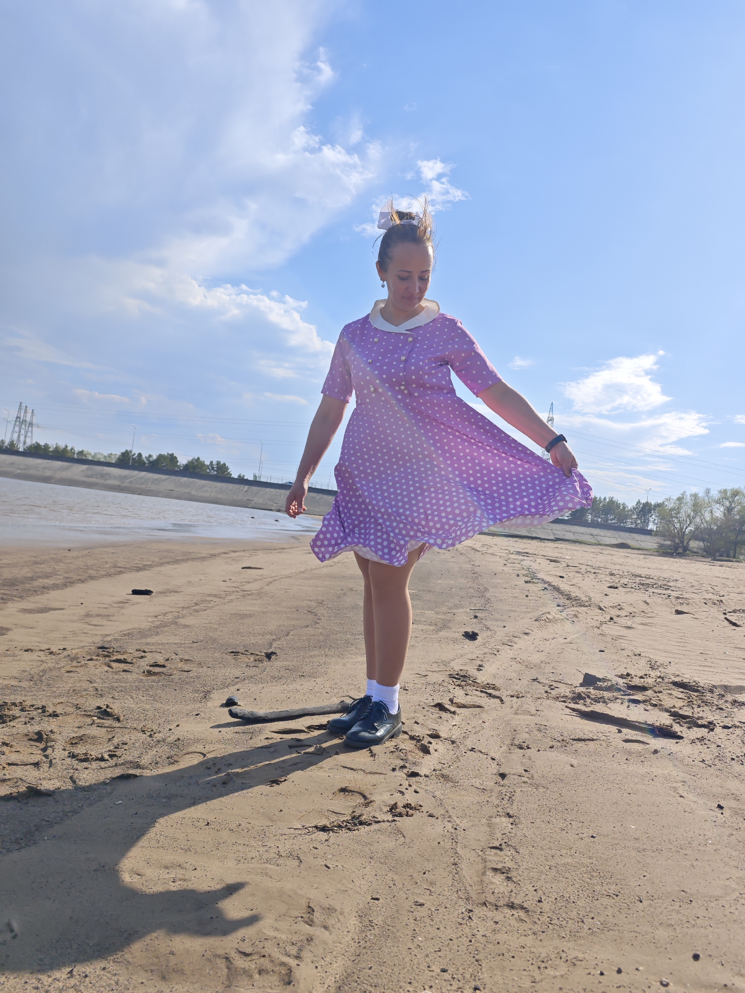 A woman is standing on a sandy beach on a sunny day. She is wearing a light purple dress with white polka dots and a white collar, white socks, and dark shoes. She is holding the edges of her dress, causing it to flare out. In the background, there is water, a concrete embankment, and trees. Utility poles and power lines are visible in the distance. The scene is in Noviy, Russia.