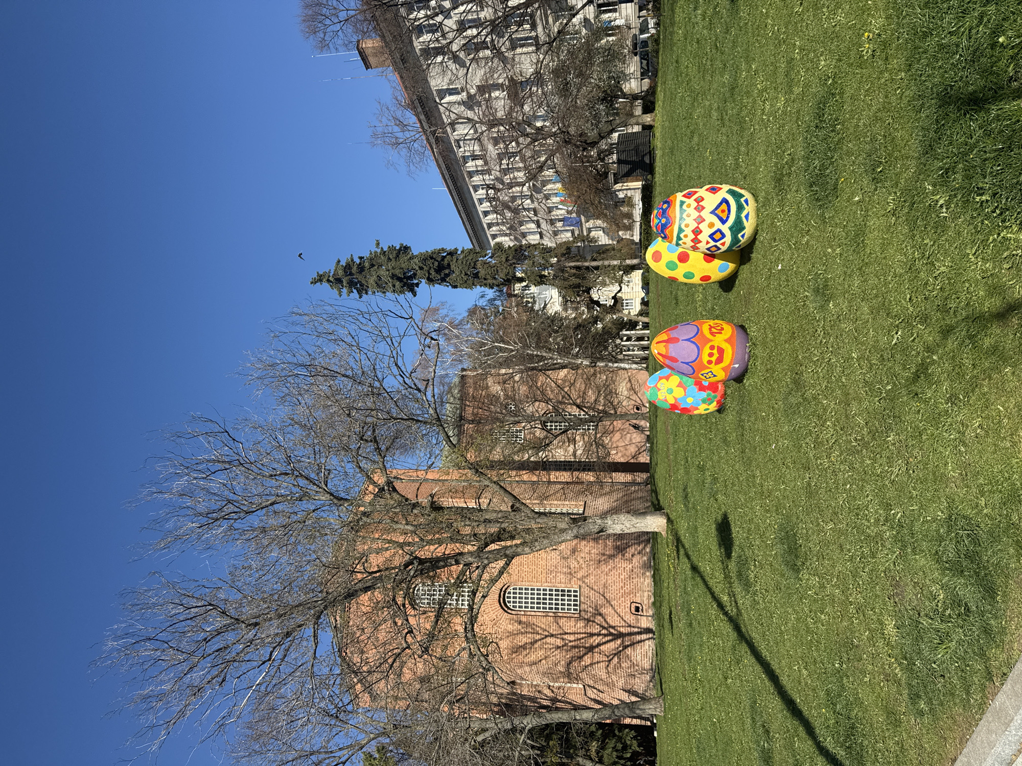 The image depicts a grassy area in Sofia, Bulgaria, with three large, decorative Easter eggs. The eggs are positioned near a brick building with arched windows and a bare tree in front of it. In the background, a large building with many windows can be seen under a clear blue sky. A bird is visible in the sky. A street light pole casts a shadow across the grass.