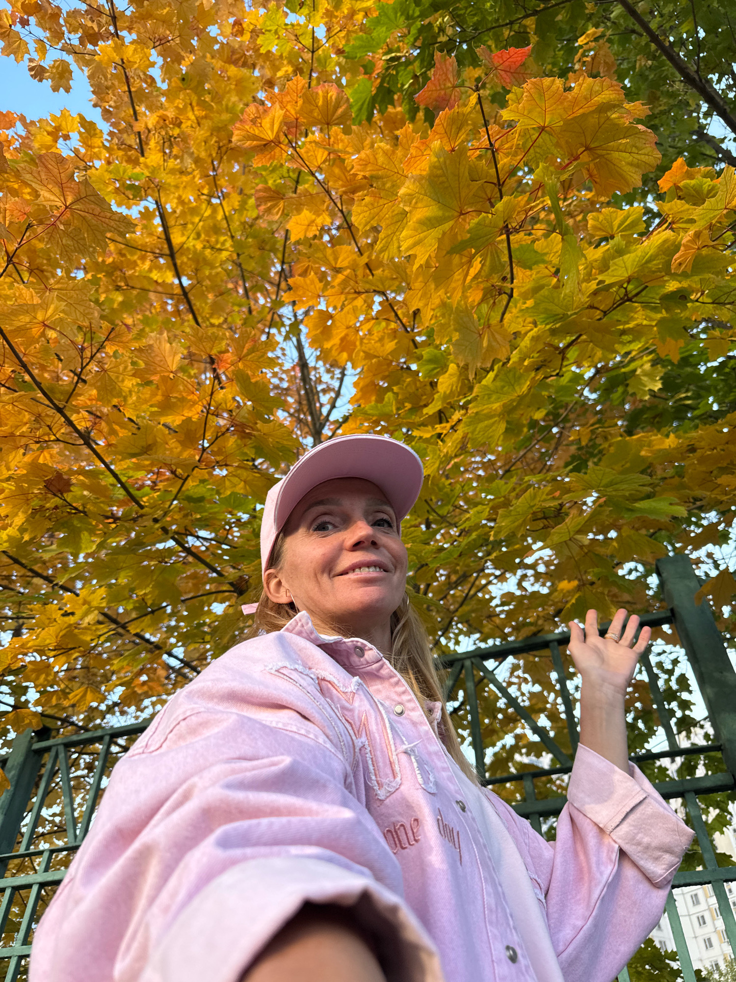 A blonde woman wearing a pink baseball cap and a light pink denim jacket poses for a selfie outdoors in front of a tree with bright yellow and green autumn leaves. She is looking towards the camera and smiling, with her right hand raised. A dark green metal fence is visible behind her, and in the distance, buildings can be seen. The setting appears to be a park or a street with trees in a residential area of Милицейский посёлок, Russia.